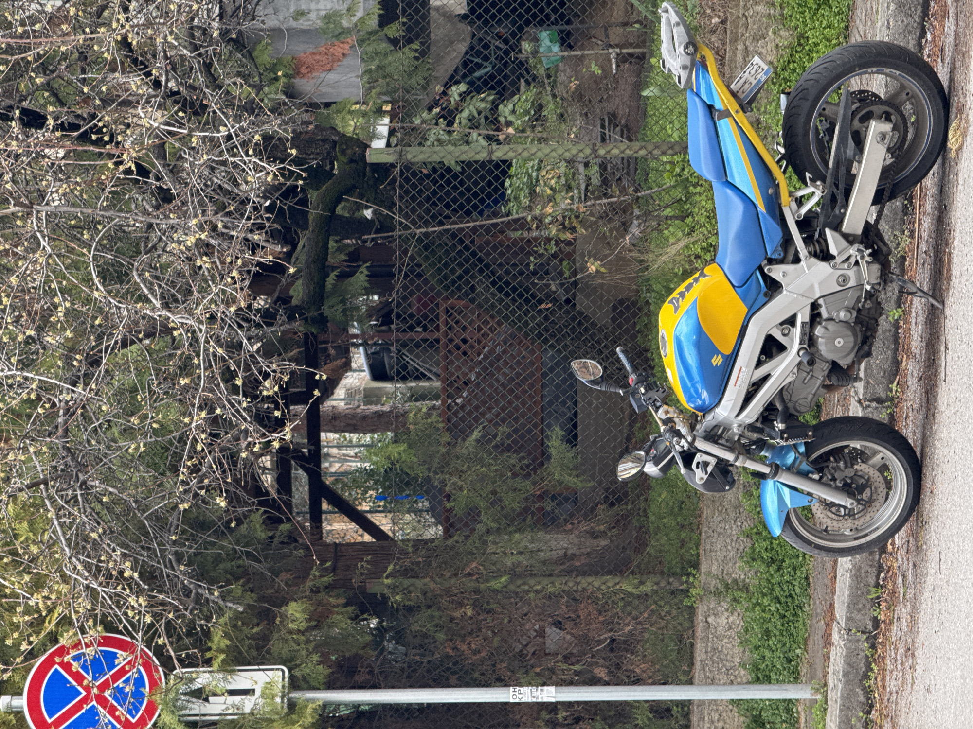 The image depicts a blue and yellow Suzuki motorcycle parked on a paved surface next to a curb, likely a road. The motorcycle is positioned in front of a wire fence, behind which is a partially obscured residential or commercial area with trees and assorted materials. To the left of the motorcycle, a leafless tree stands near a "No Parking" sign. The license plate on the motorcycle reads "CE 2853". Based on the Cyrillic alphabet used on the license plate, the location is likely in Bulgaria, and given the presence of well-maintained infrastructure and relatively dense foliage, it is plausible the image was taken in a city. A possible location is Sofia, Bulgaria.