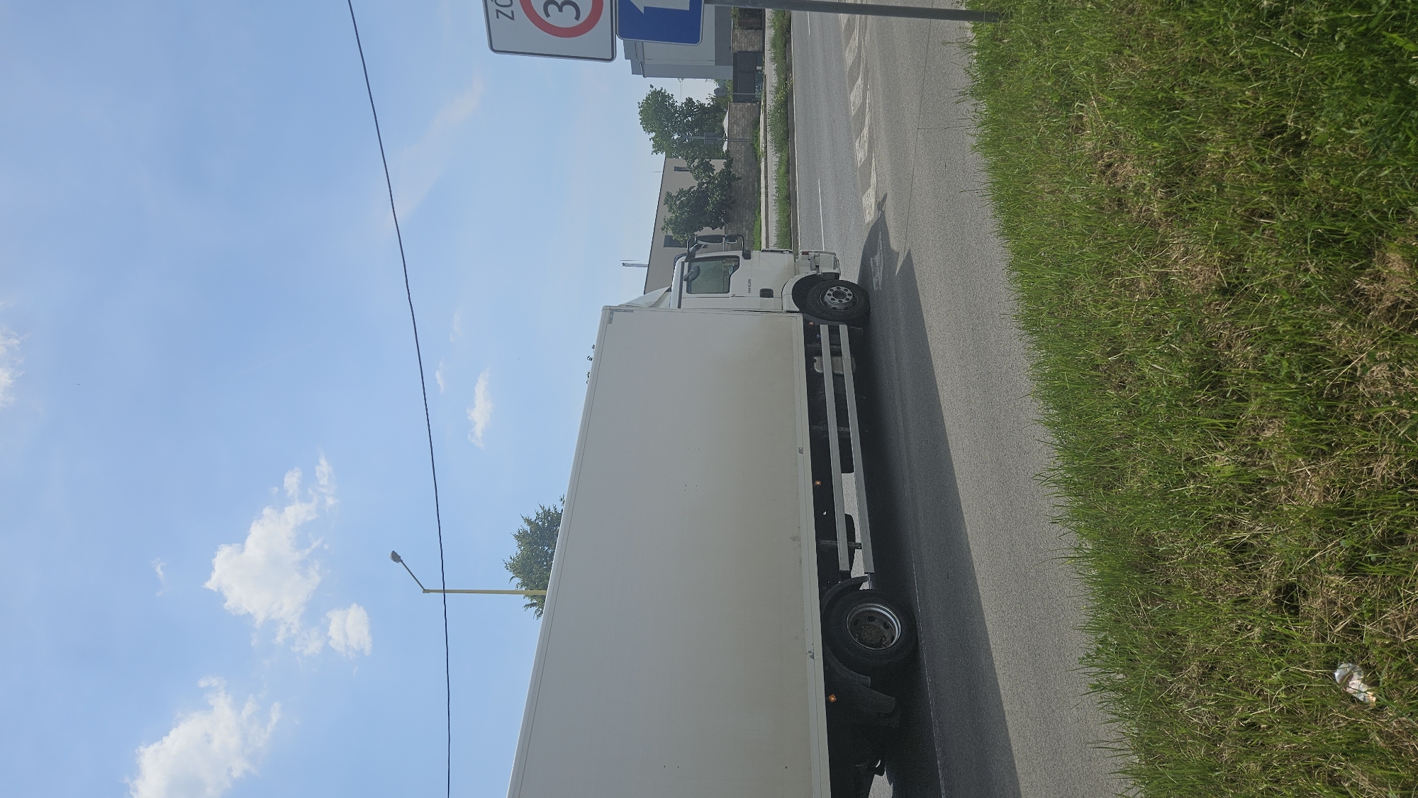 A white box truck is driving on a road in Prešov, Slovakia. The truck is in motion, indicated by its position and the slight blur on the wheels. The sky is blue with scattered white clouds. A lamppost is visible in the background, as well as some buildings and trees. A traffic sign indicating a speed limit of 30 is visible on the right side of the image. The foreground shows a grassy embankment.