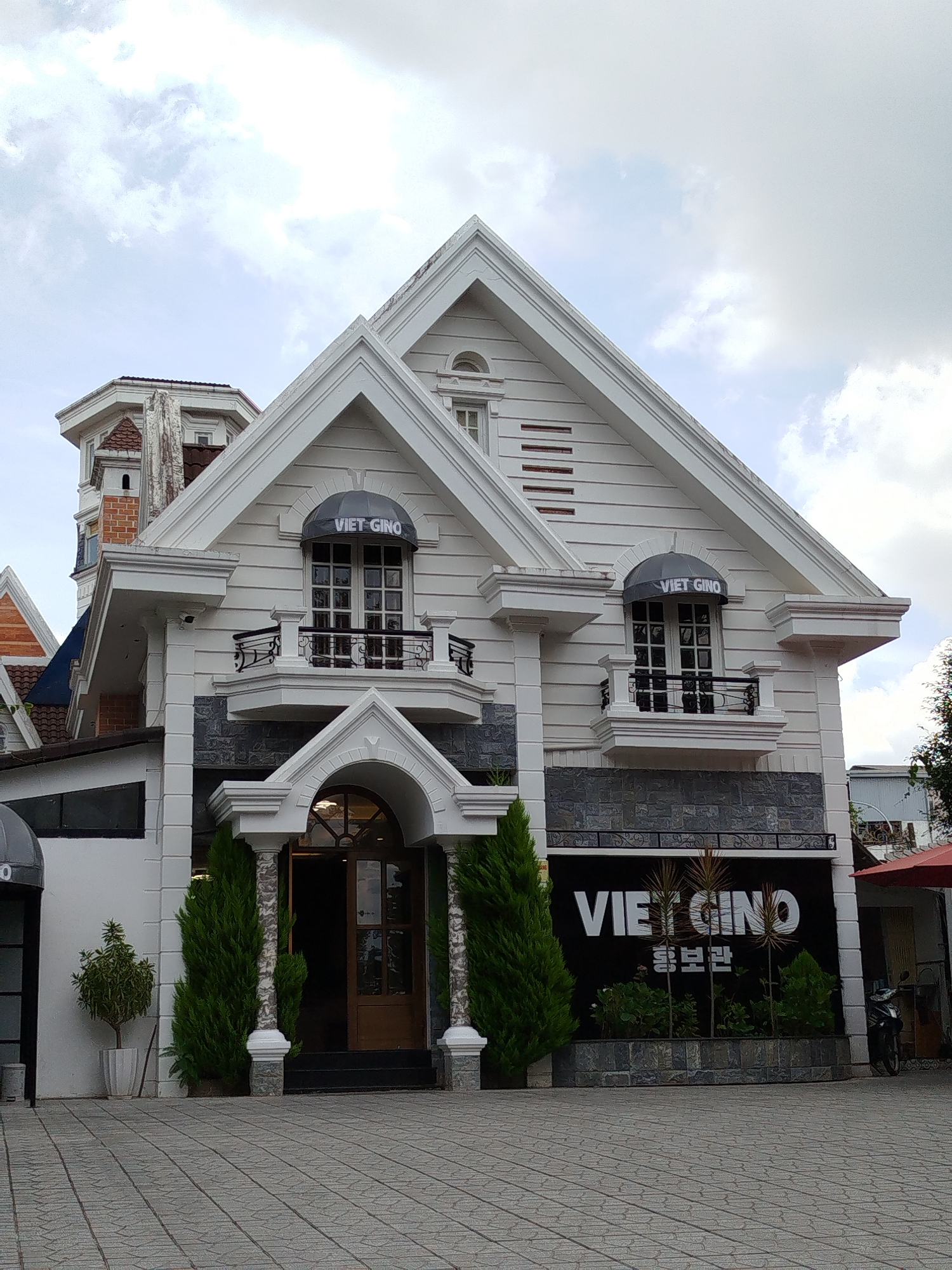 This is a medium shot of the Viet GINO building in Da Lat, Vietnam, on a cloudy day. The building is a white, two-story structure with a steeply pitched roof, architectural details like cornices and decorative trim, and dark grey stone accents on the lower level. The front entrance features an arched doorway with a wooden door and a decorative pediment above it. Two large windows with black awnings are visible on the second floor, each displaying the "VIET GINO" logo. Below these windows, small balconies with black wrought-iron railings extend outwards. A large black sign with "VIET GINO" in white lettering and Korean text below it is mounted on the lower facade, set against the stone. In front of the sign are several small green plants. To the left of the entrance, a small potted plant sits in a white planter. Two tall, slender cypress trees flank the entrance, and similar trees are seen to the right of the building. The ground in front of the building is paved with light grey rectangular tiles. To the far right, a portion of a red umbrella and a parked motorcycle are visible. The overall impression is of a modern commercial building with some decorative architectural elements, likely a restaurant or shop.