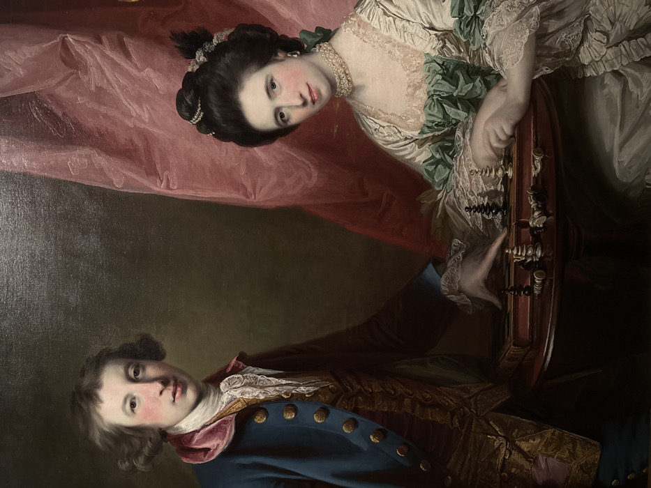 This image depicts a framed oil painting titled "The Chess Players" by Joseph Wright of Derby (circa 1780-1782), currently displayed at the Louvre Abu Dhabi, located on Saadiyat Island, Abu Dhabi, United Arab Emirates.
The painting features two individuals: a male on the left and a female on the right, both positioned in a three-quarter view, looking towards the viewer. The male, with fair skin and brown, shoulder-length curled hair, wears a blue long-sleeved jacket with gold buttons over a gold-embroidered waistcoat and a ruffled white cravat. A pink velvet collar or lapel accents his jacket. His right hand rests on a dark wooden circular table with a raised edge. The female, also fair-skinned, has dark hair styled in an elaborate updo adorned with pearls. She wears a cream-colored gown with a low neckline revealing a peach/pink undergarment, green ruffled trim, and white lace cuffs. A multi-strand pearl choker and pearl drop earrings accessorize her attire. Her hands are positioned on the table; her left hand appears poised over a chess piece.
Between the two figures, on the table, an open chessboard is visible, with numerous black and white chess pieces. Some pieces are standing upright, while others are lying scattered across the board and table surface, indicating a game in progress or recently concluded. The background consists of a dark, plain wall on the left, transitioning to a large, draped section of rich pink or reddish fabric on the right. Both figures exhibit a neutral to slightly contemplative expression.