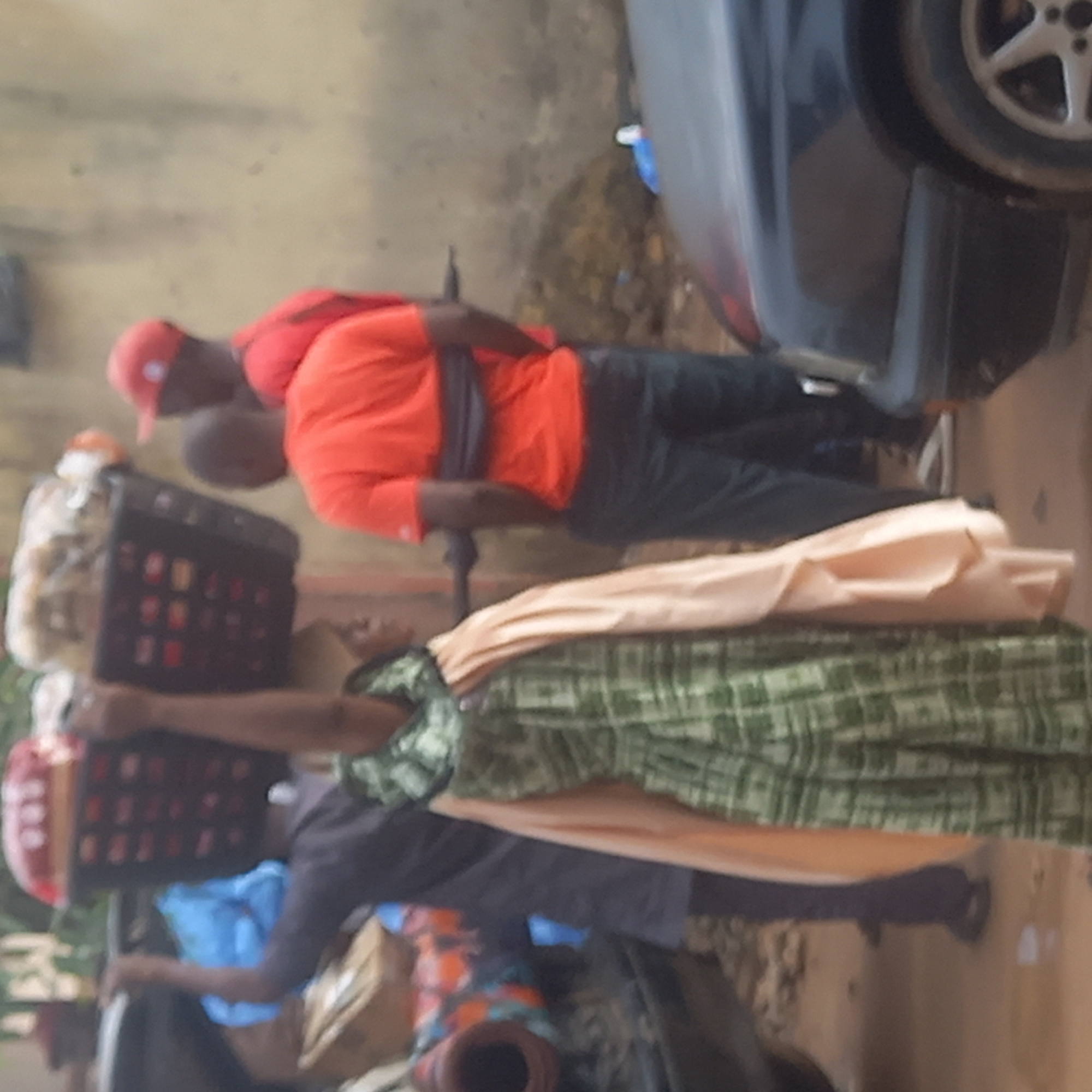 In Dadi Riba, Nigeria, two individuals are situated near a vehicle. One person is wearing a red cap and a red shirt, while the other individual is carrying baskets and draped with fabric. The surrounding environment includes what appears to be a building wall and various objects.