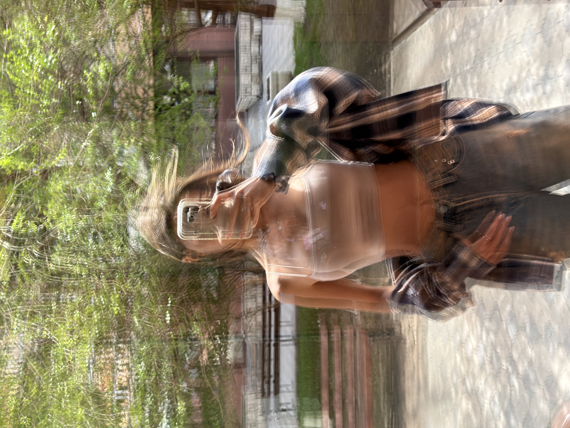 A motion-blurred photograph shows a person taking a selfie with a smartphone in Krasnoyarsk, Russia. The person is wearing a white tank top, jeans, and a plaid flannel shirt, which appears to be draped over their shoulders. Their hair is partially obscuring their face due to the motion blur. The background includes green foliage of a tree and a building with windows. The location appears to be outdoors, on a walkway or sidewalk. The environment is sunny and bright.