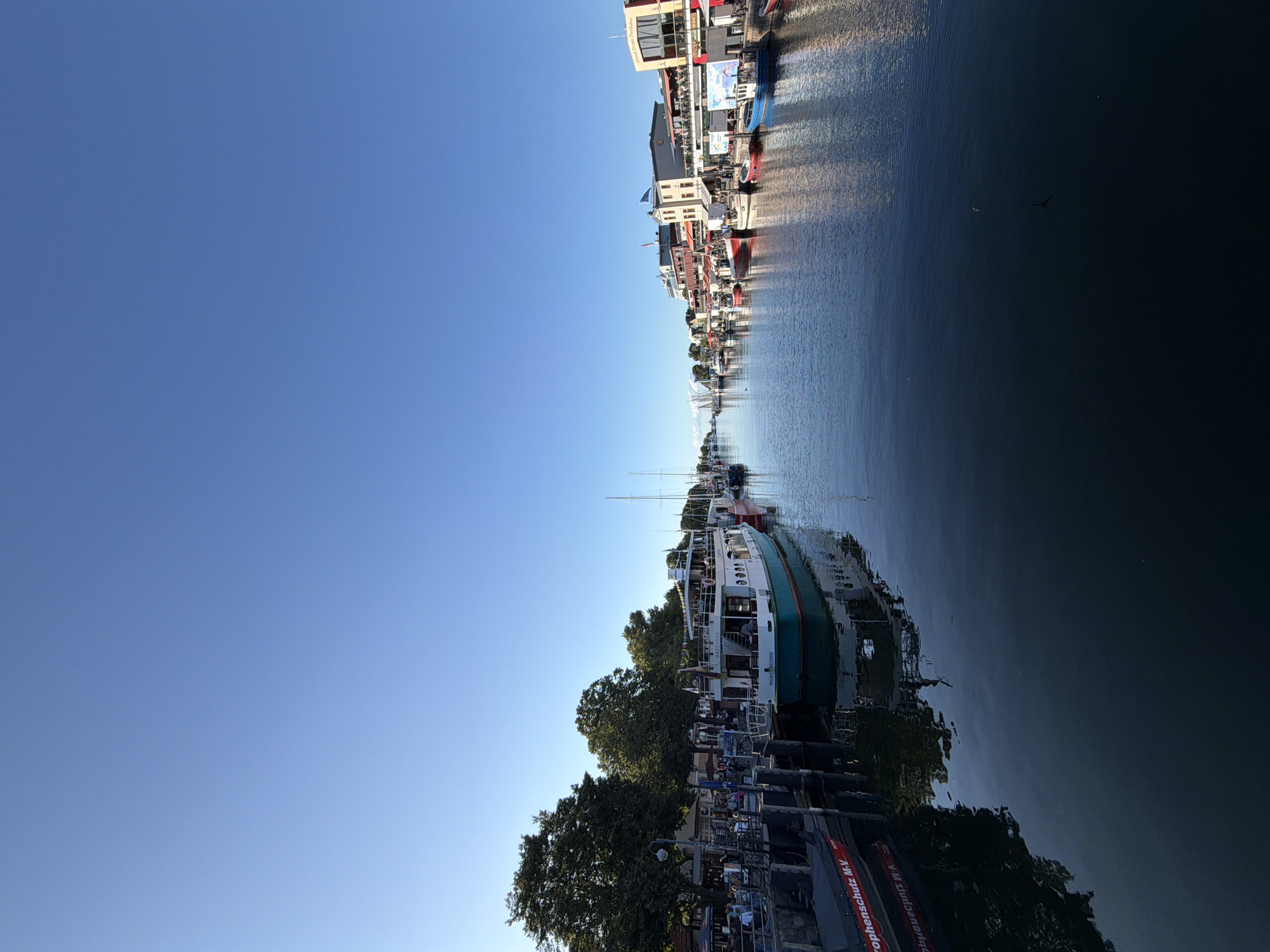 The image depicts a canal in Rostock, Germany, likely in the city harbor area, lined with boats and buildings. A clear blue sky dominates the upper portion of the frame. On the left side, a docked boat with red lettering that reads "rophenschutz M-V" is visible. Next to it, a larger white and teal boat is moored, with people seated at tables on the adjacent pier, suggesting an outdoor seating area for a restaurant or cafe. Further along the pier, more boats are docked, and buildings with multiple stories, some featuring balconies and awnings, line the canal. The water reflects the sky and the surrounding structures, creating a peaceful scene. Several other boats, including sailboats, are visible further down the canal, extending into the distance. The overall impression is of a lively, scenic waterfront in a German coastal city.