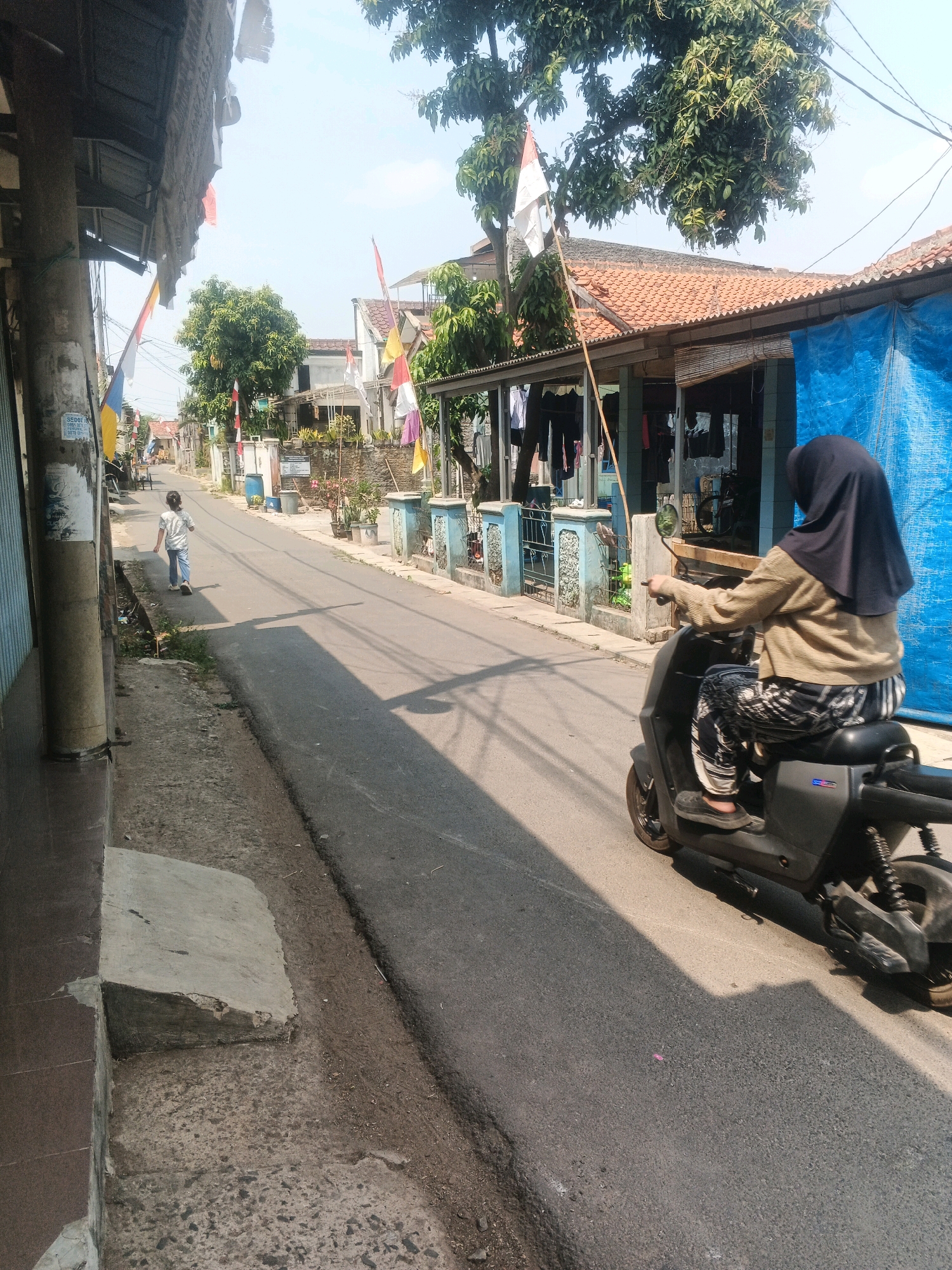 The image depicts a street scene in Kadu, Indonesia. In the foreground, a person wearing a black hijab and a beige sweater is riding a dark-colored electric scooter. They are wearing patterned pants and dark shoes. To the left of the scooter, a young child in a light-colored t-shirt and blue jeans is walking away from the camera down the road. The street is paved and appears to be in a residential area, with houses and buildings lining both sides. Some houses have tiled roofs, and one building is covered with a large blue tarp. Several Indonesian flags are visible on poles in front of the houses. Laundry is hanging on a line outside one of the buildings. The ground beside the road consists of a concrete walkway and a dirt shoulder. Sunlight casts shadows across the street. The overall impression is of a typical day in a neighborhood.