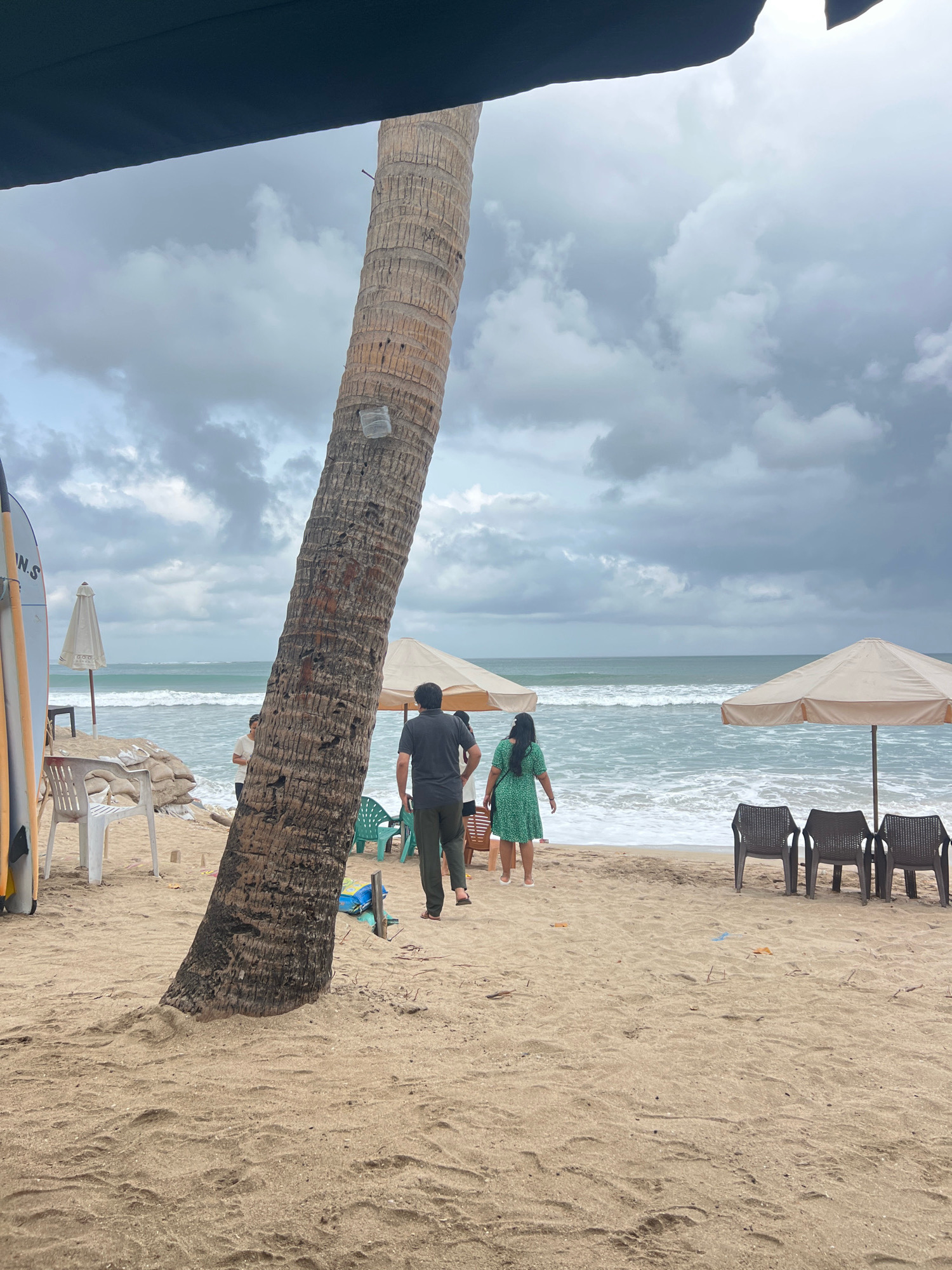 A group of people are walking on a sandy beach in Kuta, Indonesia, towards the ocean. A large palm tree is in the foreground, leaning to the left. Two beach umbrellas are visible further down the beach, with chairs underneath them. The sky is cloudy, and the ocean has moderate waves.