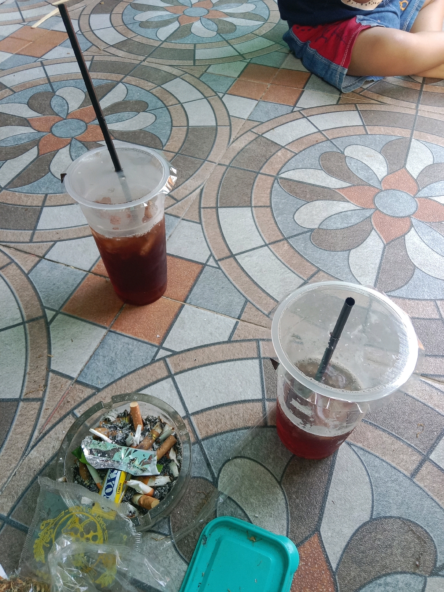 A high-angle shot shows two disposable plastic cups with dark beverages and straws, an ashtray overflowing with cigarette butts, and a child's legs in a denim skirt with red trim. The objects are arranged on a tiled floor with a floral pattern in Gambarsari, Indonesia. The tiles are in shades of beige, gray, white, and rust, arranged in a circular, flower-like design. One cup, closer to the foreground, contains a dark liquid with ice and a black straw. To the left of this cup is a clear glass ashtray filled with cigarette butts, ash, and some crumpled foil packaging. A clear plastic bag with yellow writing is partially visible in the lower left corner. The second cup, further back and to the right, also contains a dark liquid with ice and a black straw. In the upper right corner, a child's legs are visible, clad in a red skirt and denim shorts. The scene suggests a casual, outdoor setting.