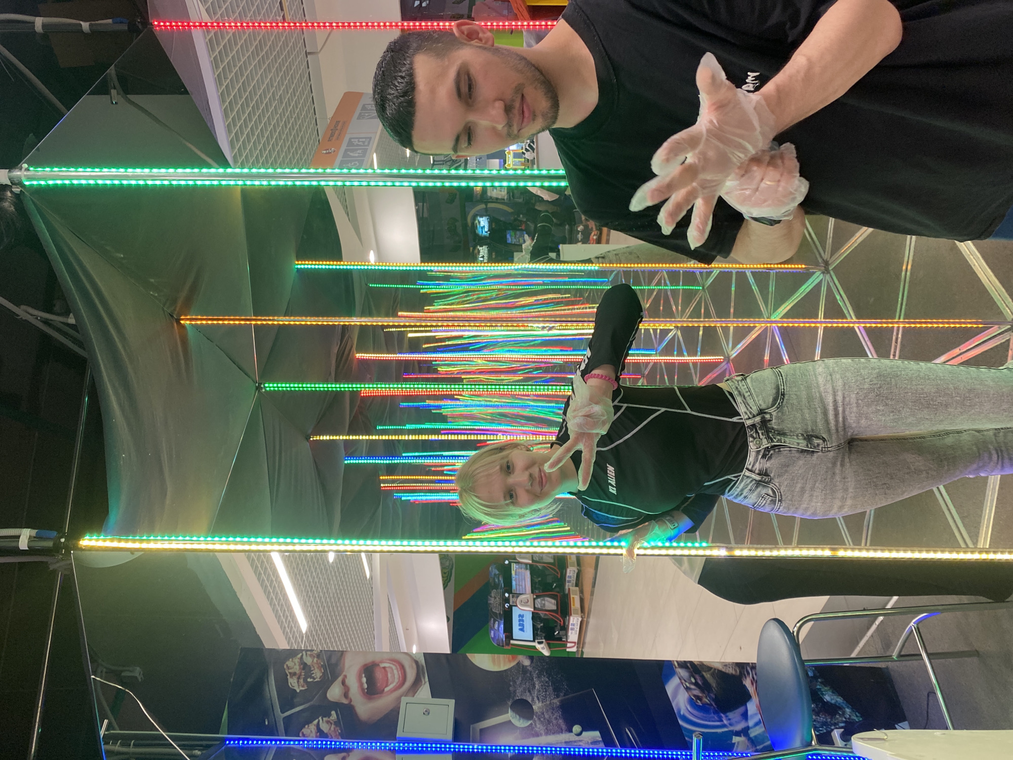 Two people are pictured inside a mirror maze. A man with short dark hair is on the top, wearing a black T-shirt and transparent plastic gloves. A young woman with blonde hair is on the bottom, wearing a black long-sleeved shirt, gray jeans, and transparent gloves. She is making a peace sign with her gloved hand. The maze is lined with mirrors and illuminated by horizontal rows of colored LED lights. The reflection in the mirrors reveals a variety of surfaces, including advertisements, equipment, and structural elements of the location. The chair and table are visible in the image, potentially of the location or part of the maze. The setting appears to be an entertainment venue in the city of Chaikovsky, Russia.