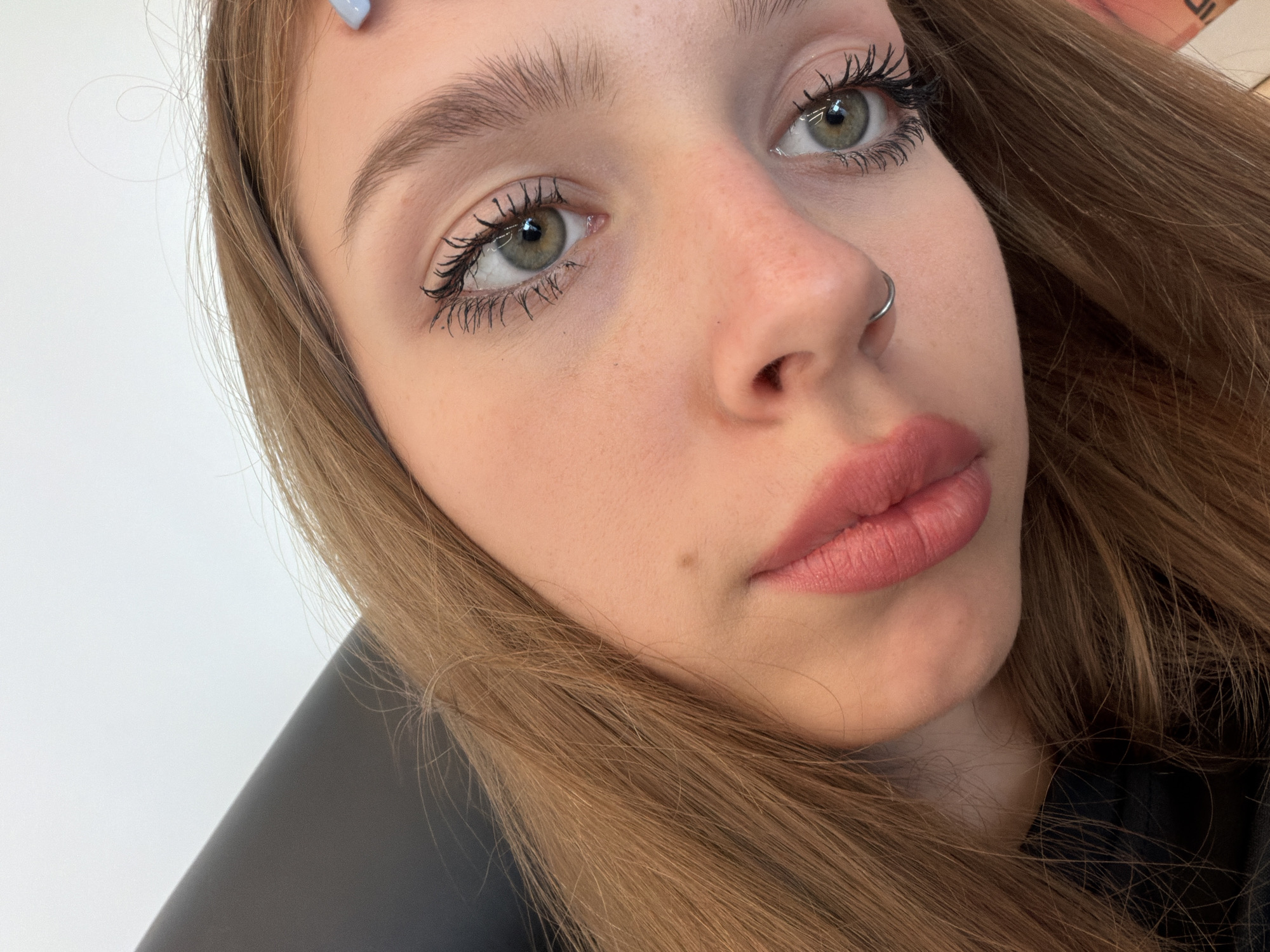 This media file is a close-up, low-angle selfie of a young woman with light brown hair and light-colored eyes. She is looking directly at the camera. The woman has on makeup, including mascara and lipstick. She is wearing a nose ring. Her hair is long and falls around her face and shoulders. The background is mostly a plain white wall, with a subtle shadow cast across it. There are no visible text elements in the image. The lighting appears to be soft, suggesting an indoor setting during the daytime, but the exact time of day or weather conditions cannot be determined. The image captures a neutral expression.