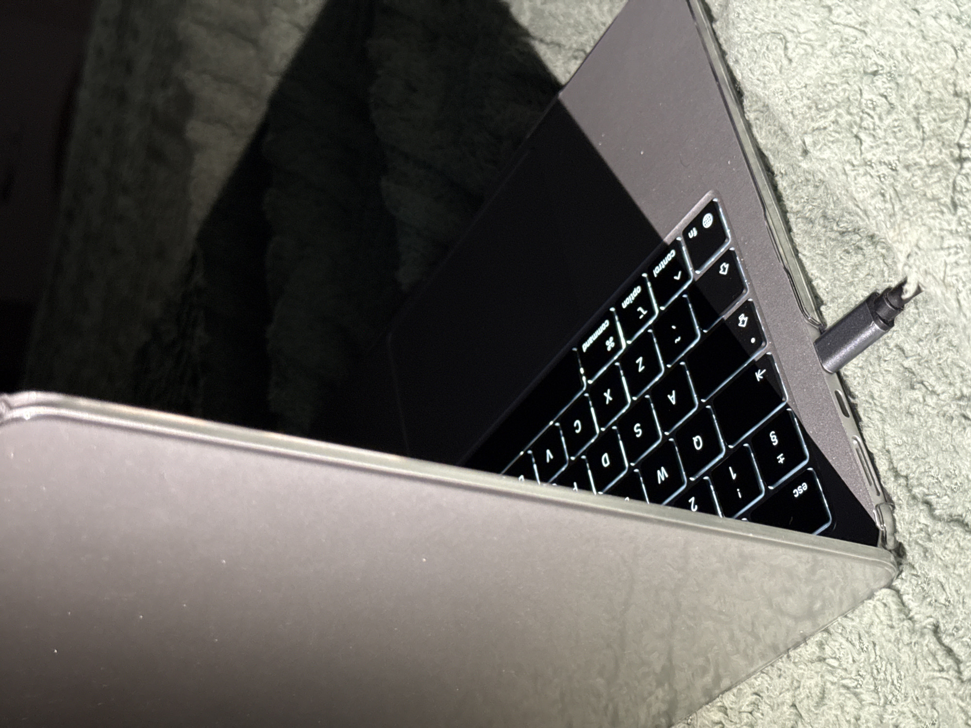 The image displays a close-up, angled view of a laptop, likely a MacBook, with its lid partially open. The laptop is placed on a textured, light green surface, possibly a blanket or rug. The keyboard is illuminated with white backlighting, revealing individual keys. A USB-C cable is plugged into the side of the laptop, with its connector visible. The lighting in the scene is dramatic, with strong shadows cast across the laptop and the surrounding surface, suggesting either natural light or a focused artificial light source. The laptop appears to be in a domestic setting, given the soft furnishings. The image does not contain any information or context that would allow for its location to be specified within the city of Slavyani, Bulgaria.