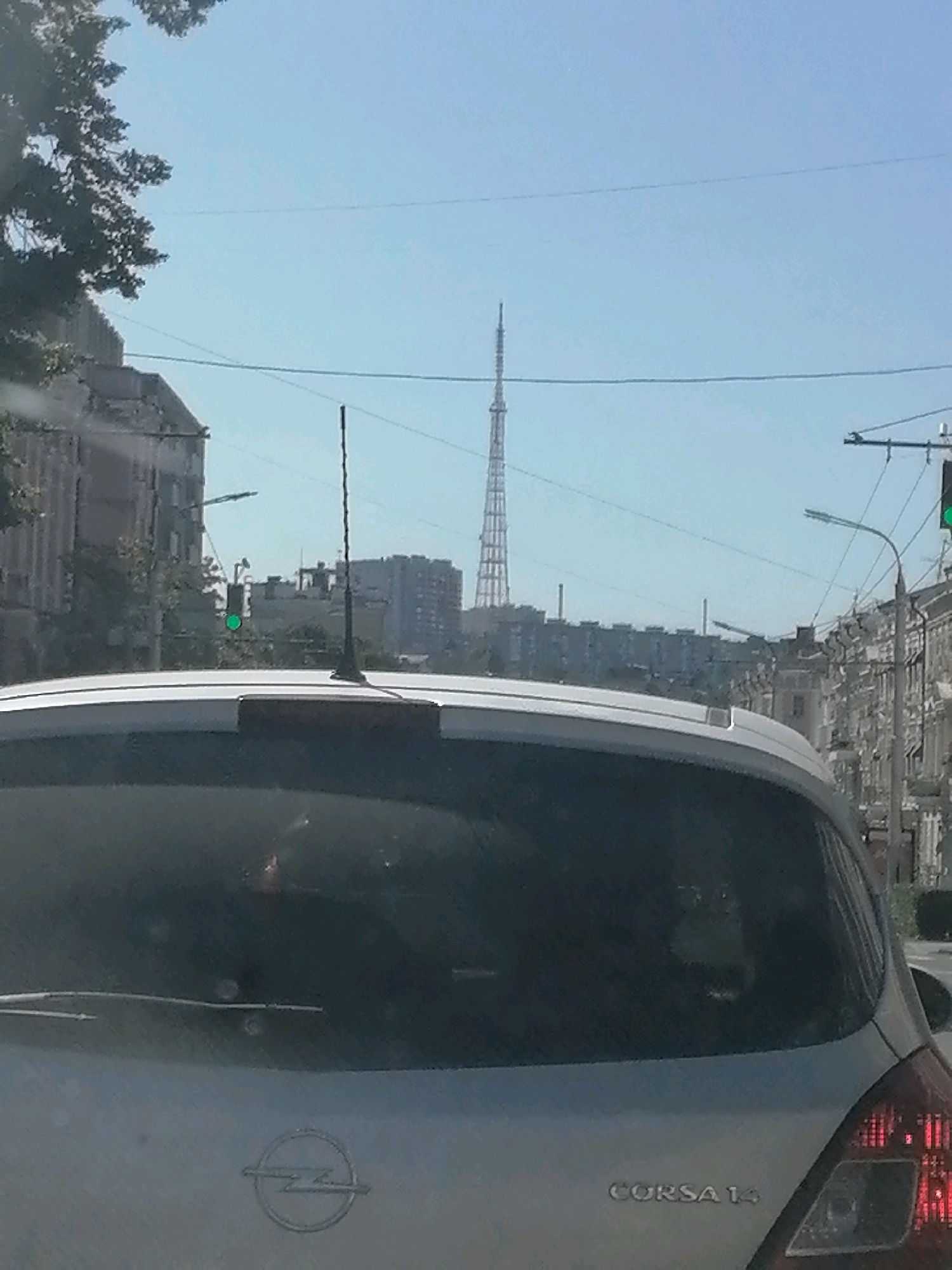 The image, taken from inside a vehicle, looks out towards a city street with buildings and a tall radio tower in the distance. The sky is clear and blue. In the foreground, the rear windshield of the vehicle is visible, with the Opel logo and "CORSA 14" badging on the tailgate. A traffic light showing green is visible on the left side of the frame. The scene appears to be a sunny day in Rostov-on-Don, Russia, likely on a street with a view of the city's landmarks.