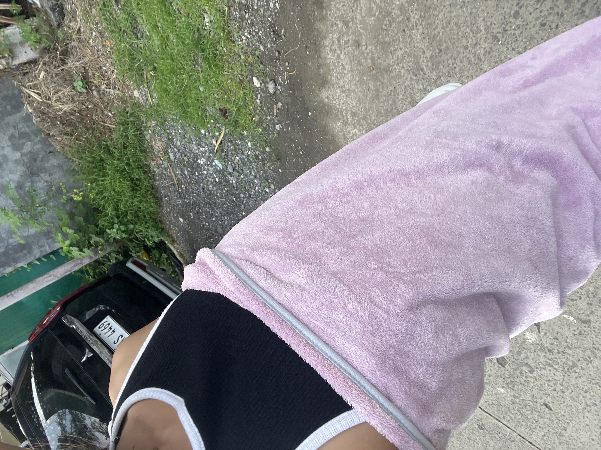 A person is standing outdoors, partially wrapped in a pink towel. They are wearing a black tank top with white trim. In the background, a black vehicle with a license plate that reads "S 4469" is parked near a wall and some greenery. The ground is a mixture of concrete and gravel. The scene appears to be in an urban or suburban area, possibly a residential street in Davao City, Philippines, given the car's license plate format and the surrounding vegetation.