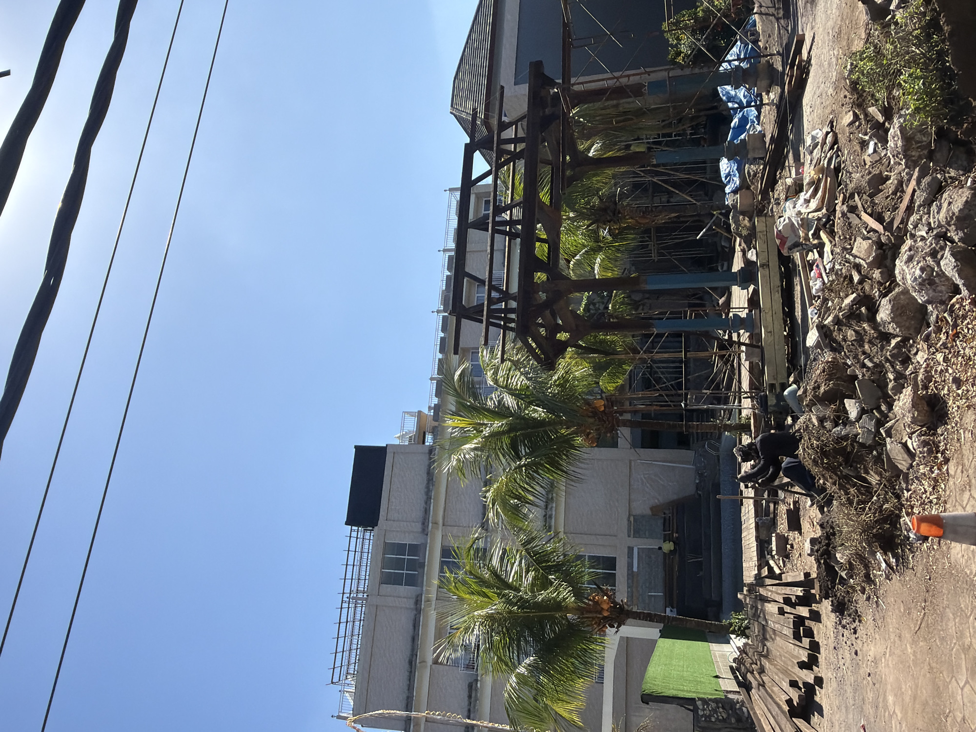 The image depicts a construction site in Kuta Selatan, Indonesia, under a clear blue sky. In the foreground, there is a pile of rubble and construction debris, including rocks, wood, and dirt. Several pieces of lumber are stacked to the left. In the center of the frame, a wooden gazebo structure is under construction, supported by blue pillars and scaffolding. Two people are visible on the construction site: one is sitting on the ground and appears to be working with tools, while another is standing in the background near a pool. Behind the gazebo, two palm trees with coconuts are prominent. Further in the background, a multi-story building, possibly a hotel or resort, is under construction. Power lines are visible stretching across the top of the image. The overall scene suggests an active construction zone with ongoing building and landscaping work.