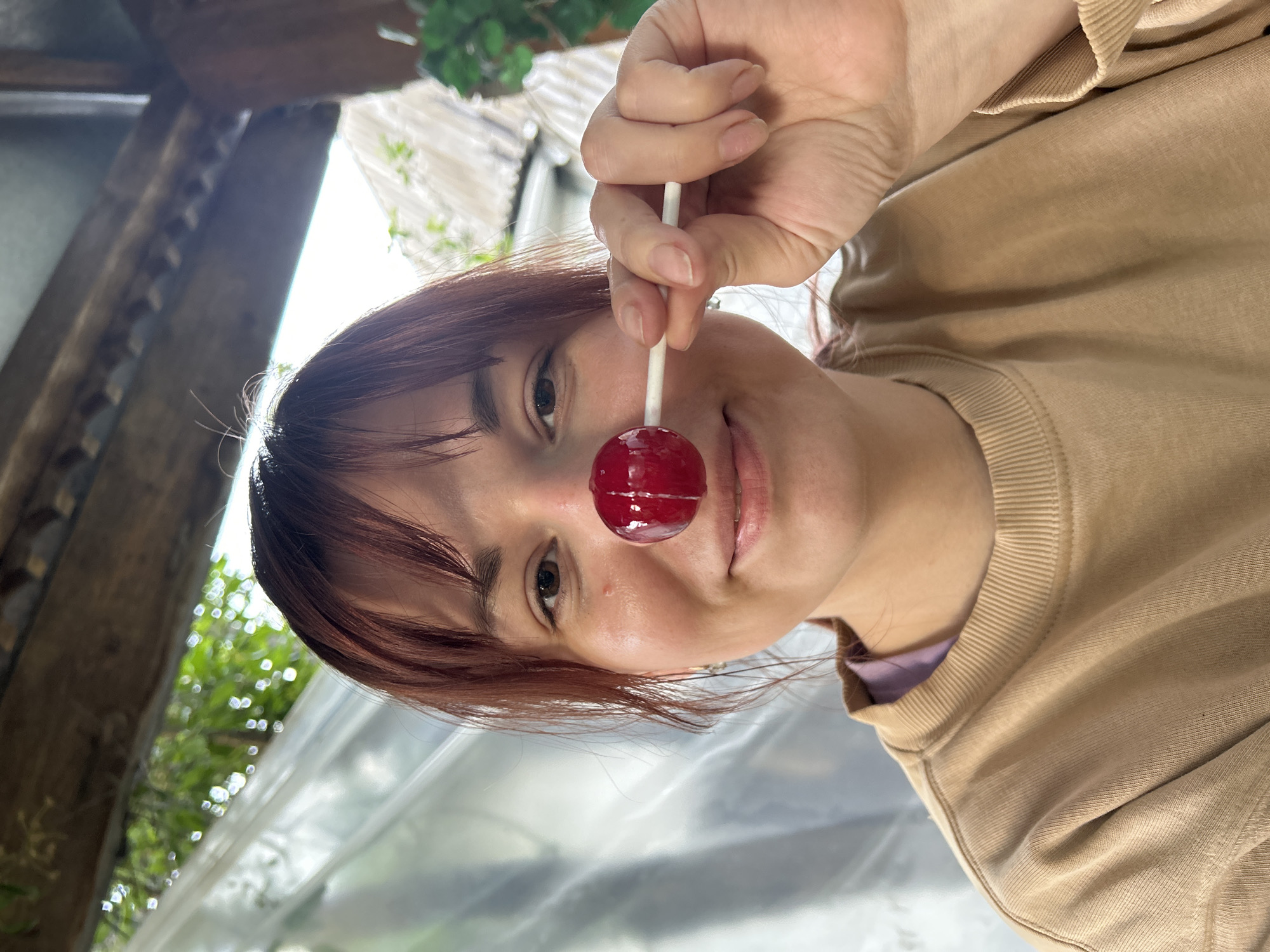 A young woman with auburn hair and bangs is holding a red lollipop in front of her nose. She's wearing a beige sweatshirt with a purple undershirt visible at the neck. The background shows a wooden structure, likely a building or enclosure, along with green foliage and a bright, slightly overcast sky. The exact location in the city of Фоки, Russia, cannot be determined with the visual information available.