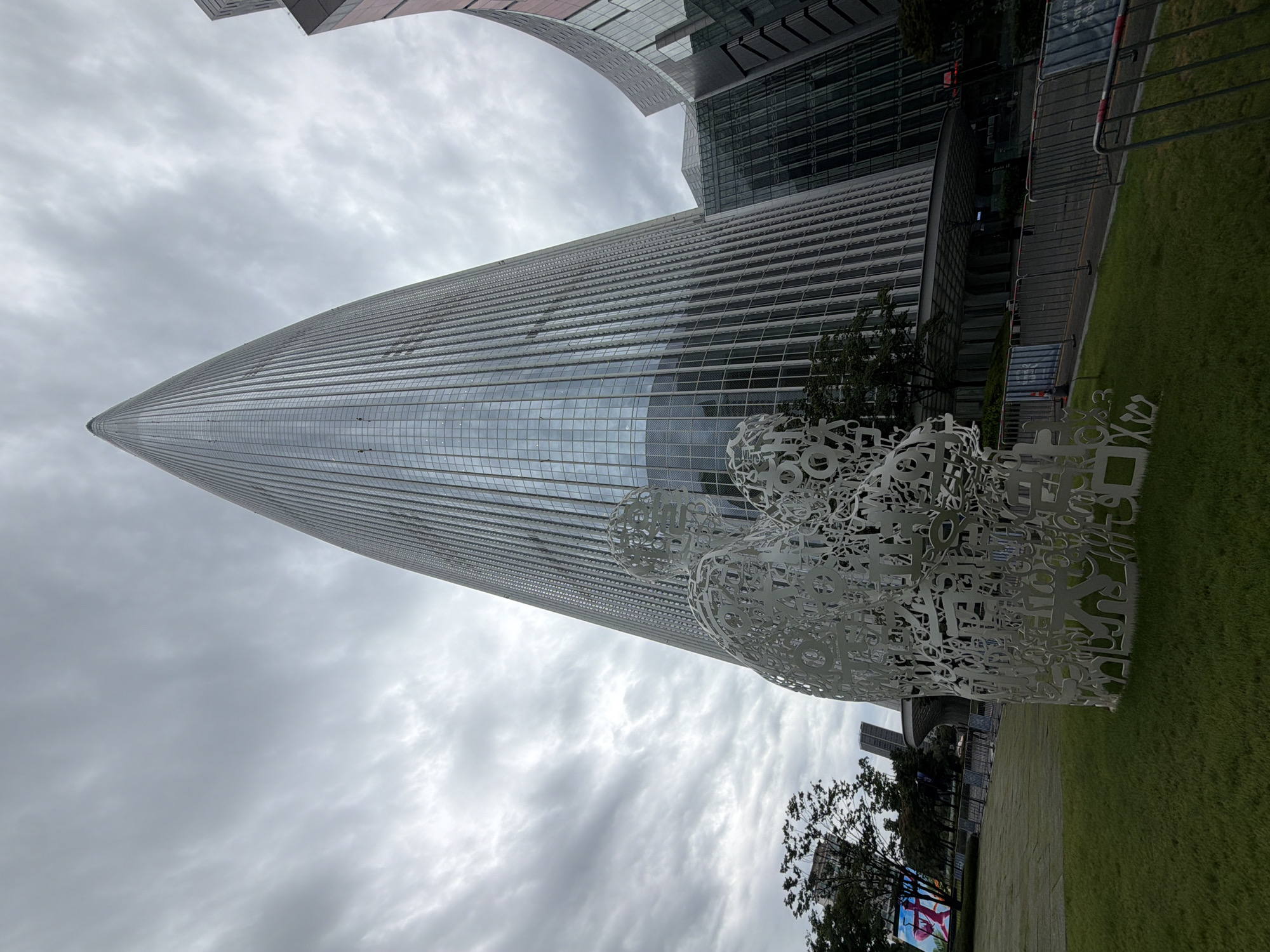 The image captures a view of Lotte World Tower in Seoul, South Korea, on an overcast day. In the foreground, a large, intricate white sculpture made of abstract shapes and what appear to be letters and numbers is situated on a patch of well-maintained green grass. Behind the sculpture, the imposing Lotte World Tower dominates the skyline, its glass facade reflecting the cloudy sky. To the right of the tower, other modern buildings with curved glass exteriors are visible, adding to the urban landscape. A metal fence separates the grassy area from a walkway or road in front of the buildings. Trees are sparsely planted around the area, and in the background, other buildings and a large digital billboard can be seen. The overall impression is one of modern architecture set against a dramatic, cloudy sky. There are no discernible individuals in the image, and the focus is on the architectural elements and the sculpture.
