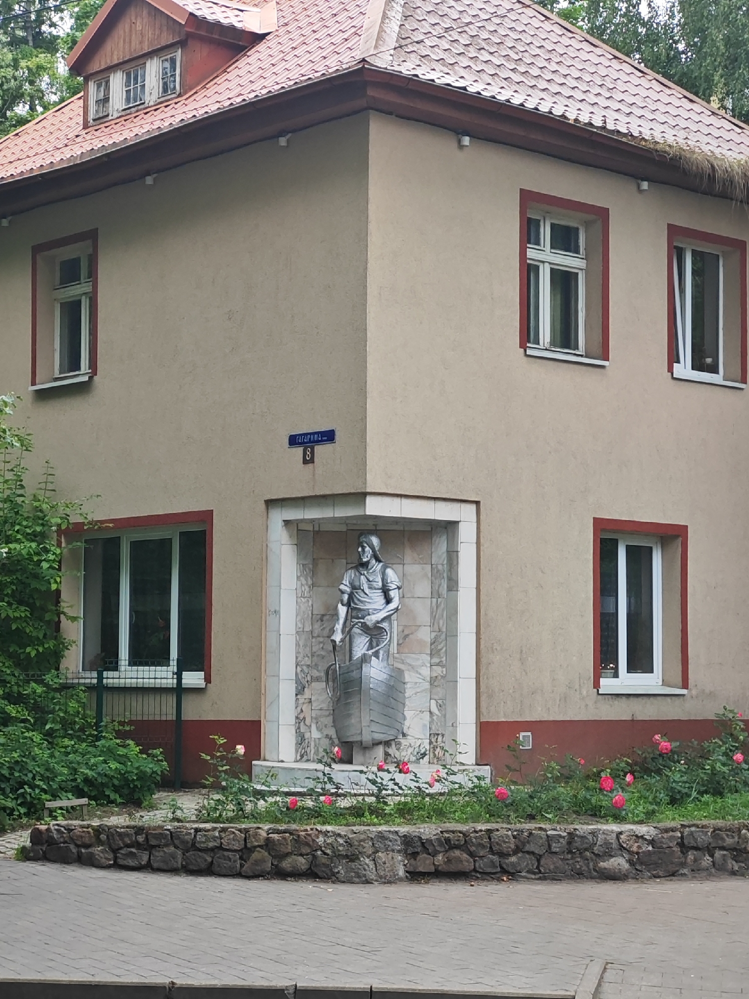 The image shows a side view of a building with a red-tiled roof. The building is light beige with red trim around the windows. On the corner of the building, there is a niche with a silver-colored statue of a person dressed in what appears to be work clothes, holding a tool and a cylindrical object. The statue is set against a backdrop of marbled tiles.
In front of the statue, there is a small garden with green bushes and several pink roses in bloom. The garden is bordered by a low wall made of rough stones. Beyond the garden, a paved walkway is visible, suggesting an outdoor public space.
Above the niche, a blue street sign with white lettering is mounted on the building wall. The visible text on the sign appears to be "Гагарина" (Gagarina) and the number "8".
The scene is lit by natural light, suggesting it is daytime. The weather appears to be pleasant, as evidenced by the blooming roses and the foliage on the trees in the background. The overall atmosphere is calm and perhaps historical, given the presence of the statue and the street name. The location is identified as Svetlogorsk, Russia.