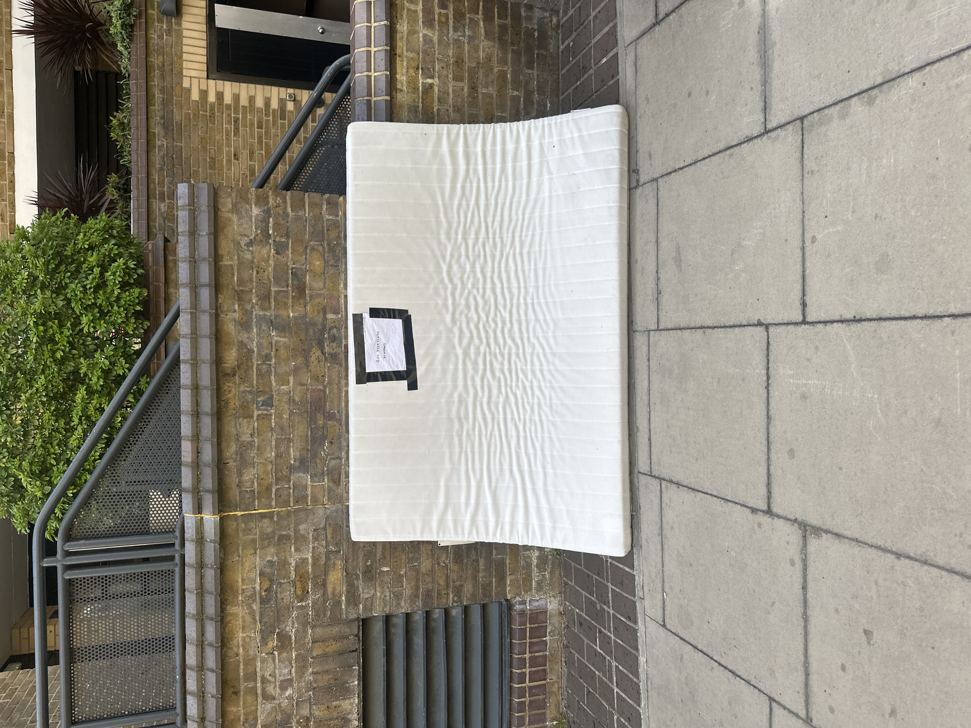 A white mattress is leaning against a brick wall on a paved sidewalk. The mattress has a dark sticker attached. Adjacent to the brick wall is a building entrance with metal railings and greenery. The setting appears to be an urban environment.