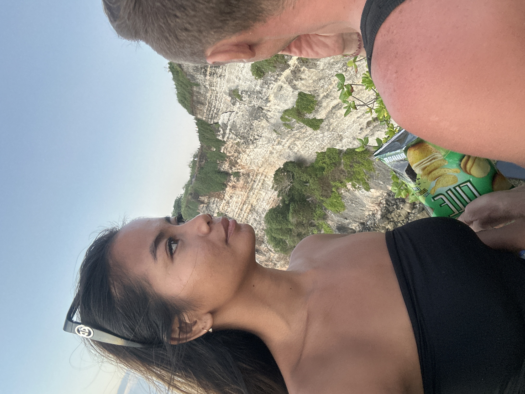 In Kuta Selatan, Indonesia, a woman with dark hair, wearing sunglasses on her head and a black strapless top, looks to her right. She is standing with a man, whose back is to the camera, holding a bag of chips. They are positioned on a cliff overlooking the ocean, with steep, rocky cliffs covered in greenery in the background. The sky is clear and blue.