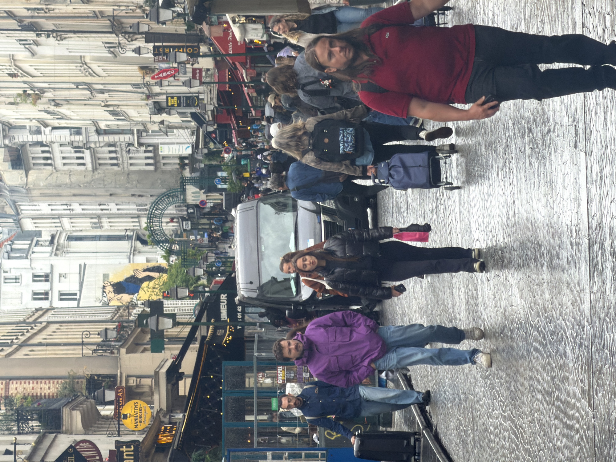 This is an image of a street in Paris, France, likely in the Montorgueil neighborhood. The street appears crowded with pedestrians. A car is visible amongst the crowd. Numerous storefronts and signs are present along the street, including "MANHATTN'S -BURGERS- Montorgueil," "UMEUR," and "TABAC". Several people are walking, some with luggage. The street surface appears wet, reflecting the surroundings. The buildings are typical of Parisian architecture.