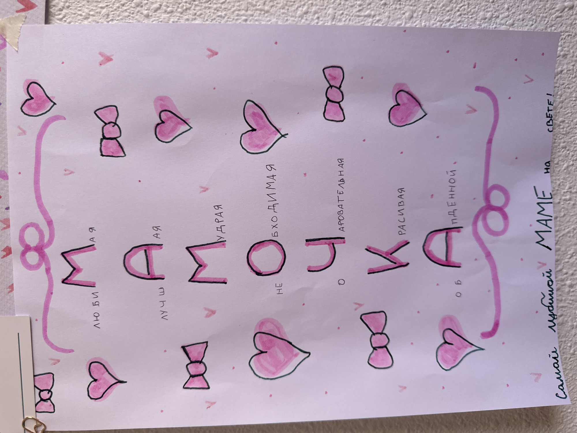 The image shows a child's drawing on white paper. The drawing is done in pink and black ink. It features the word "МАМОЧКА" (MAMOCHKA - meaning Mommy) spelled out with decorative letters. Below each letter are words in Russian that describe the mother, such as "люби" (love), "лучш" (best), "мудрая" (wise), "необходимая" (necessary), "очаровательная" (charming), "красивая" (beautiful), and "объединенной" (united). The word "Самой любимой МАМЕ на свете!" (The most beloved MOM in the world!) is written in black ink at the bottom. The drawing is decorated with pink hearts and bows scattered around the paper. The drawing is likely a gift or a message for Mother's Day. The location and other contextual details cannot be determined from the image.