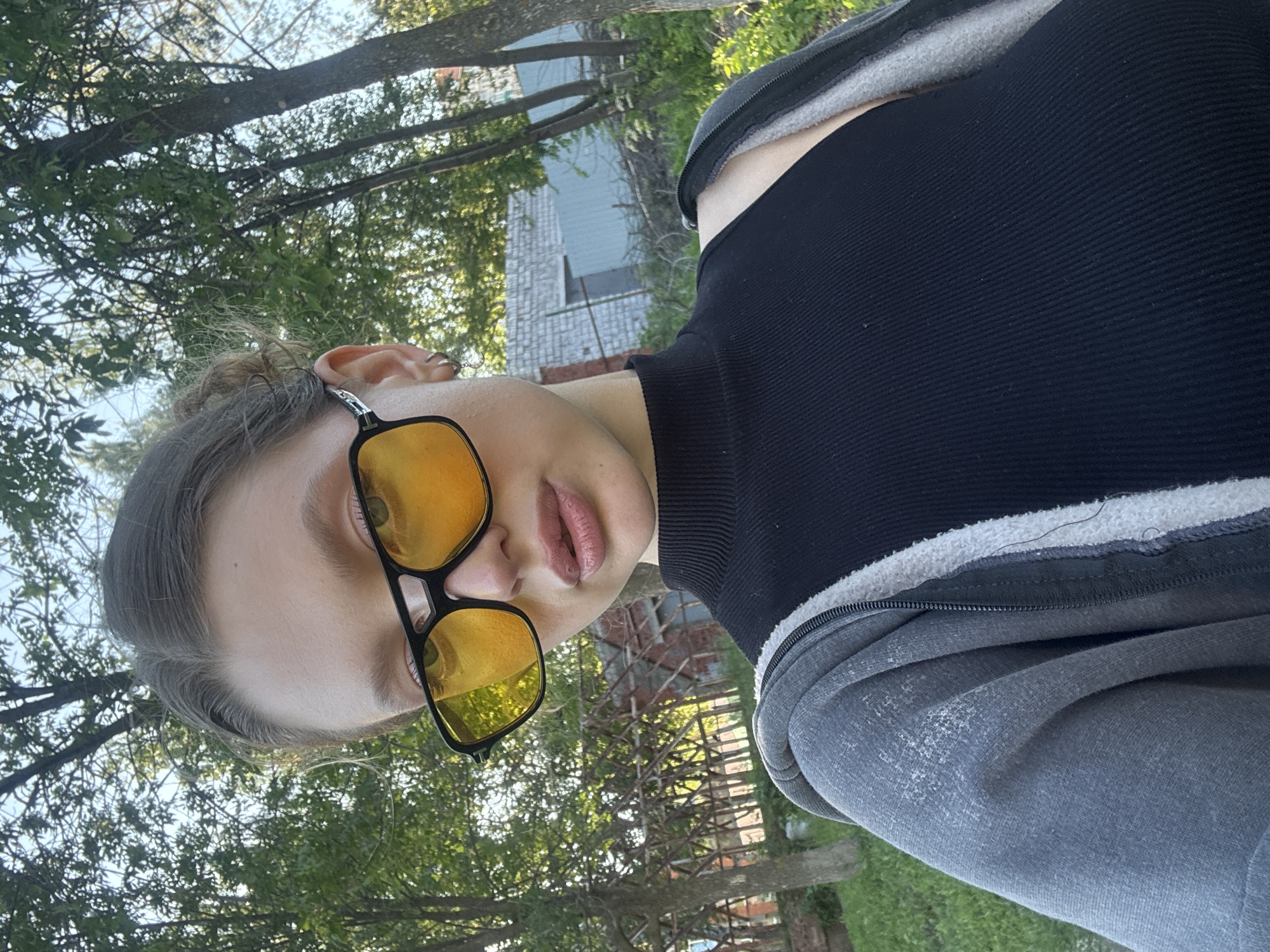 A young Caucasian woman with brown hair is taking a selfie outdoors. She is wearing a black ribbed turtleneck top and a grey hooded zip-up jacket. She has on large, dark-rimmed sunglasses with yellow lenses. Her hair is pulled back into a bun. The background suggests a residential or semi-rural area with trees, some type of building construction, and a blue corrugated metal fence. The lighting indicates it is daytime, likely late afternoon or early evening, given the warm tones.
It is not possible to determine the specific location within Izhevsk, Russia from the visual information provided in the image.