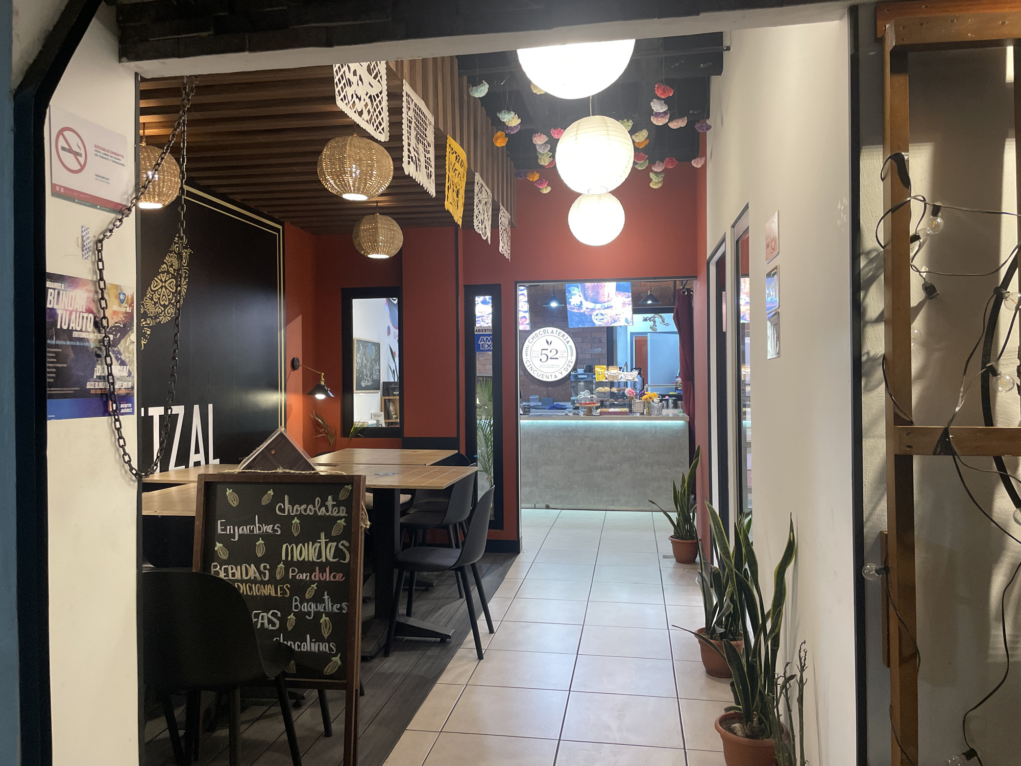 Here is a detailed description of the image:
The image shows the entrance to a chocolatier in Mexico City, Mexico, specifically "Chocolateria Cincuenta y Dos (52)". The view is from just outside the entrance, looking into the establishment.
On the left, a poster that says "BLINDAR TU AUTO BENITO JUAREZ" is partially visible. A chain hangs near the entrance. A chalkboard menu lists items such as chocolates, enjambres, molletes, pan dulce, baguettes and "chocolinas". Tables and chairs are visible in the foreground of the restaurant area.
The interior of the chocolatier is decorated with festive elements including paper cutouts, hanging spherical lamps, and colorful decorations on the ceiling. A counter with pastries is seen further inside. Potted snake plants are arranged along the right wall leading into the shop. The color scheme includes shades of orange, white and light grey. String lights decorate the right side of the entrance. The flooring is tile, and there appears to be a transition to a different flooring under the tables on the left.
The scene presents a warm, inviting atmosphere with a focus on traditional Mexican artisanal decorations and treats.