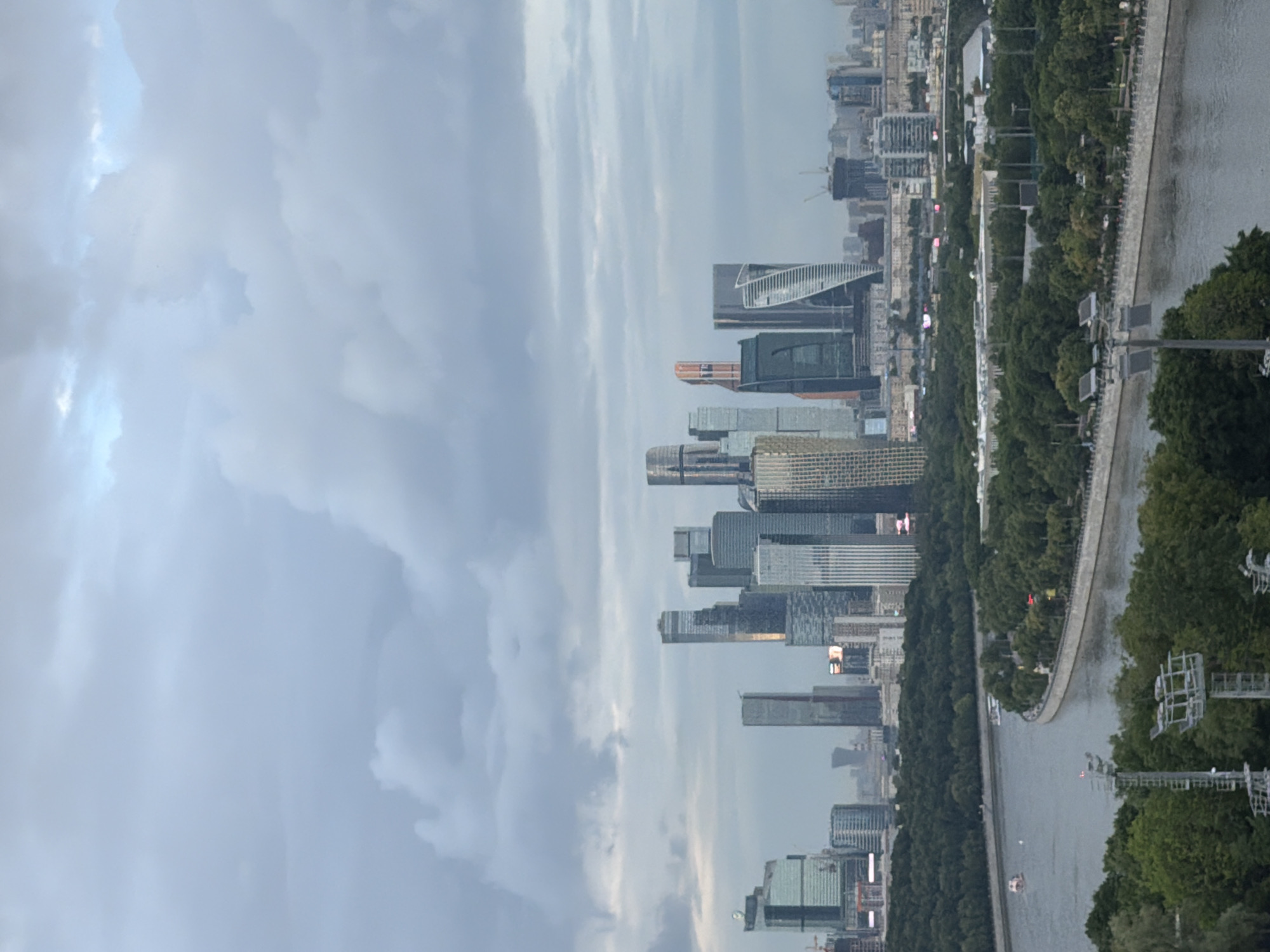 An aerial, medium-wide shot captures a city skyline dominated by modern skyscrapers, a winding river, and lush green parkland under a cloudy sky. The skyscrapers, a mix of glass and steel structures, rise prominently in the background, with some reflecting the overcast sky. A wide river curves through the midground, with a paved embankment lined by dense trees and a pathway. Several boats are visible on the river. In the foreground, parkland with more trees and some infrastructure, including lighting towers, is visible. The overall impression is that of a bustling urban environment integrated with significant green spaces.