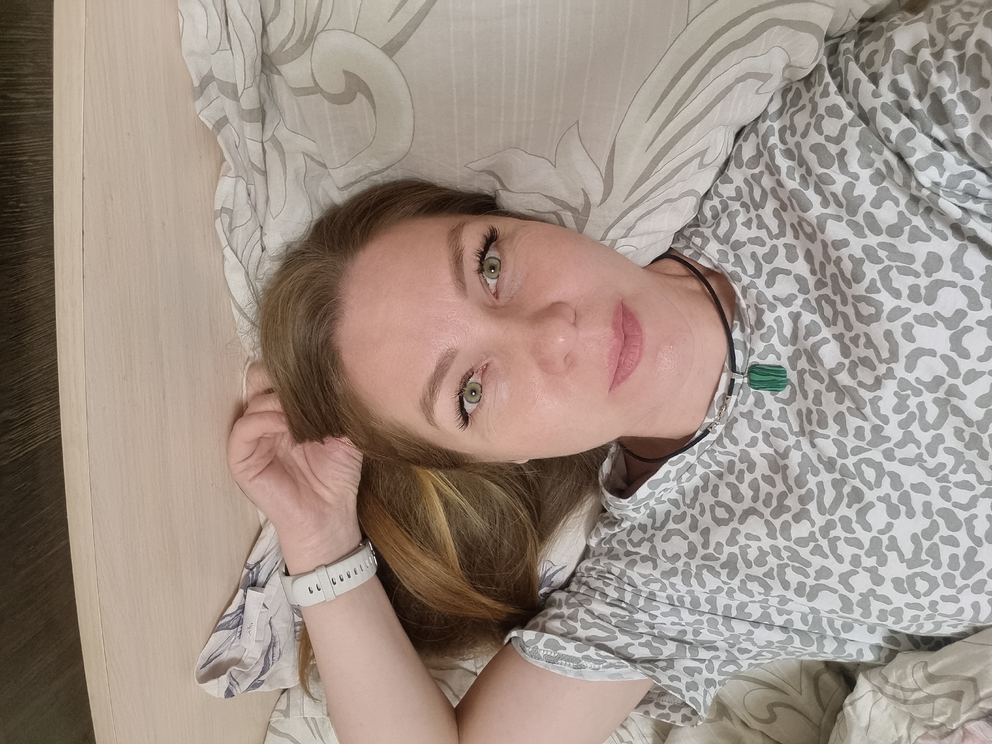 An adult woman is captured in a close-up, overhead shot, lying in bed and looking directly at the camera with a gentle, faint smile. Located in Chaikovsky, Russia, she appears relaxed in an indoor setting, likely a bedroom. Her long, light brown hair with subtle blonde highlights spreads around her head on a light-colored pillow. She has striking green eyes and well-defined eyelashes and eyebrows.
She is wearing a light-colored top with a grey, abstract animal print. Around her neck, a dark cord necklace is visible, featuring a rectangular, layered green pendant, possibly made of malachite. A white digital watch with visible perforations is worn on her left wrist.
The bedding, including the pillow and what appears to be a duvet or sheet, is a light tone with a subtle grey and white swirling or floral pattern. To the left side of the frame (viewer's right), a light wooden panel, possibly part of a headboard or wall, is visible, alongside a darker, textured surface that may be a floor or furniture. The lighting is soft and even, suggesting a bright indoor environment, likely during the day. No discernible text is clearly visible on any items, though a tiny, unreadable label is present on her clothing. The overall impression is one of calm and tranquility.