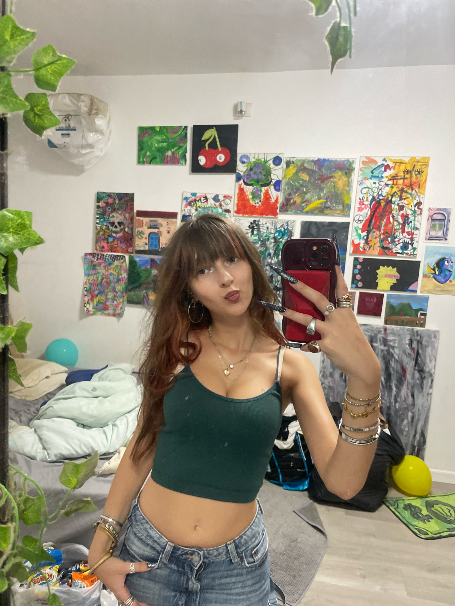 A young woman with long brown hair and bangs is taking a selfie in a room, reflected in a mirror. She is wearing a dark green crop top and distressed denim shorts. She has long, dark, pointed fingernails with a checkered pattern on some. Her right arm is raised, holding a red phone. She is wearing several rings on her fingers and bracelets on her wrist. She is also wearing a necklace with a heart pendant. Behind her, the wall is covered with numerous small, colorful paintings and artwork. To her left, a bed is visible with rumpled light-colored bedding and a light blue balloon. A large plant with green leaves hangs from the top left of the mirror frame. The floor appears to be laminate or wood. The overall impression is of a cluttered, artistic bedroom. There is no indication of Raanana, Israel.