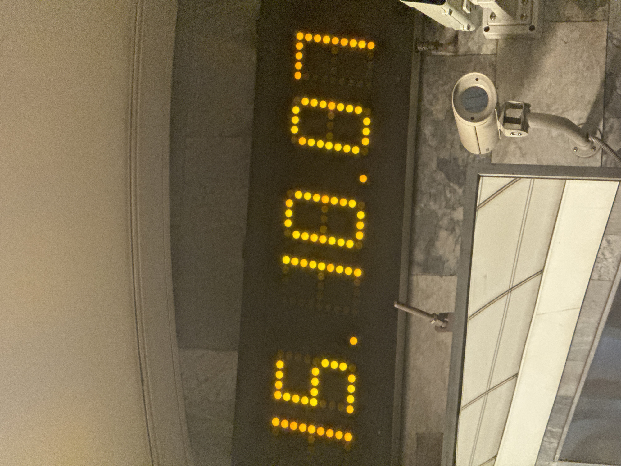 Located in Saint Petersburg, Russia, the image features a large, vertical LED digital display, showing bright yellow numbers. When oriented to be read horizontally (90 degrees clockwise rotation), the display shows "16.10.07," which could represent a date (e.g., October 7th, 2016) or a precise time (e.g., 16 hours, 10 minutes, 7 seconds).
To the right of the display, a light-colored, cylindrical security camera is mounted on a textured wall, possibly made of stone or tile. This camera is angled downwards, suggesting it monitors the area below. Above it, part of another mounting bracket or a second camera is partially visible. Below the primary camera, a geometric-patterned structure, resembling a window or architectural paneling, is seen. To the left of the digital display, a plain, light-colored wall with a subtle vertical groove adds to the structured environment.
The scene appears to be set in low light conditions, which enhances the prominence of the glowing display. There are no people visible, and no specific activity or event is explicitly taking place beyond the display showing numerical information. The setting suggests an indoor or covered outdoor public space, such as a transit station or a commercial building.