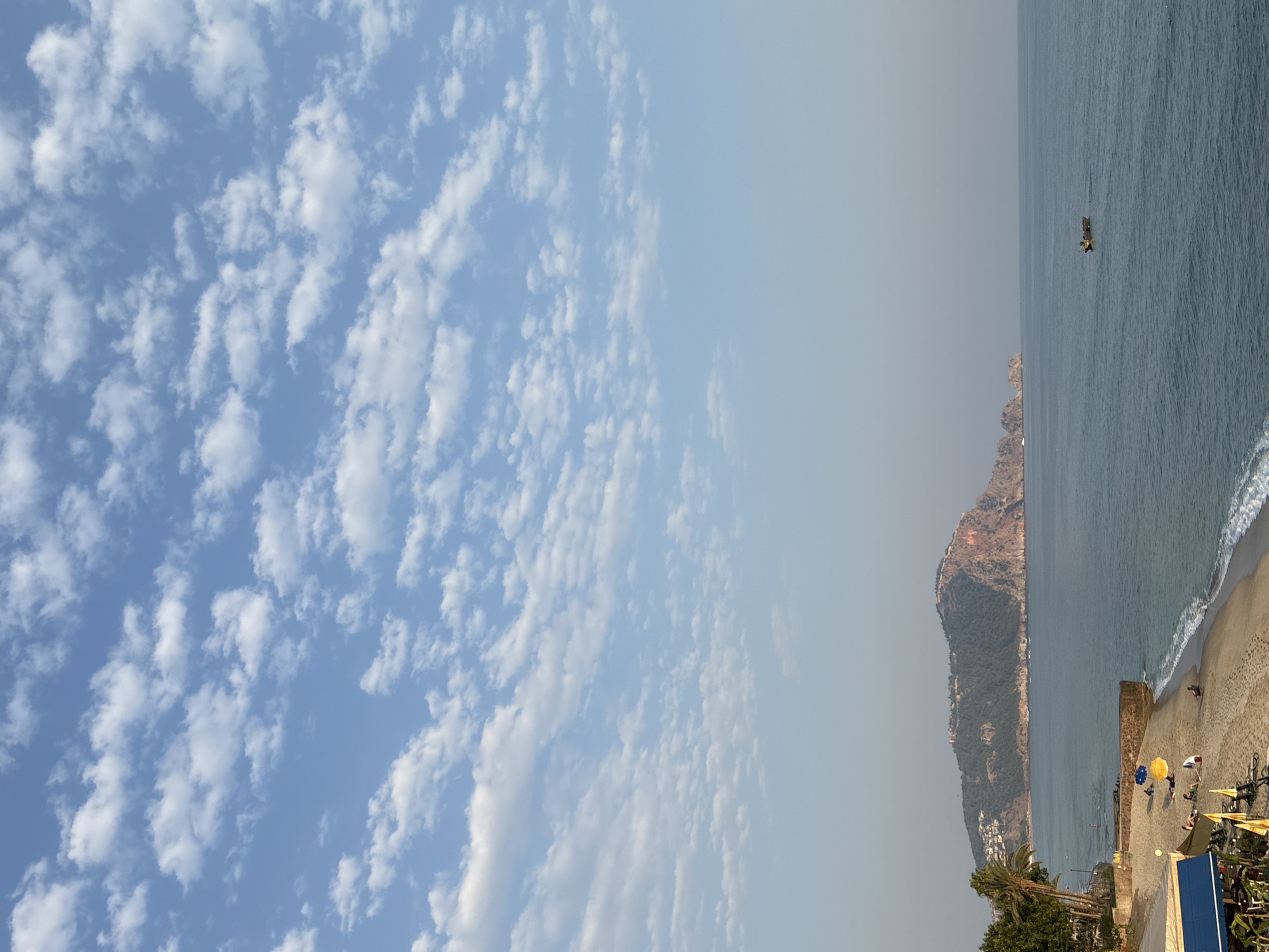 The image depicts a coastal scene in Alanya, Turkey, with a prominent rocky peninsula jutting out into the sea. A large, rugged mountain with scattered buildings and a historical fortification at its summit dominates the background. The foreground features a sandy beach with a few scattered umbrellas and people relaxing. Palm trees line the edge of the beach, and a small restaurant or cafe with blue and yellow awnings is visible on the left. The sea is a calm, deep blue, with a small wooden boat with two occupants visible in the distance. The sky is a clear, bright blue, dotted with scattered, wispy white clouds. The overall atmosphere is serene and tranquil.