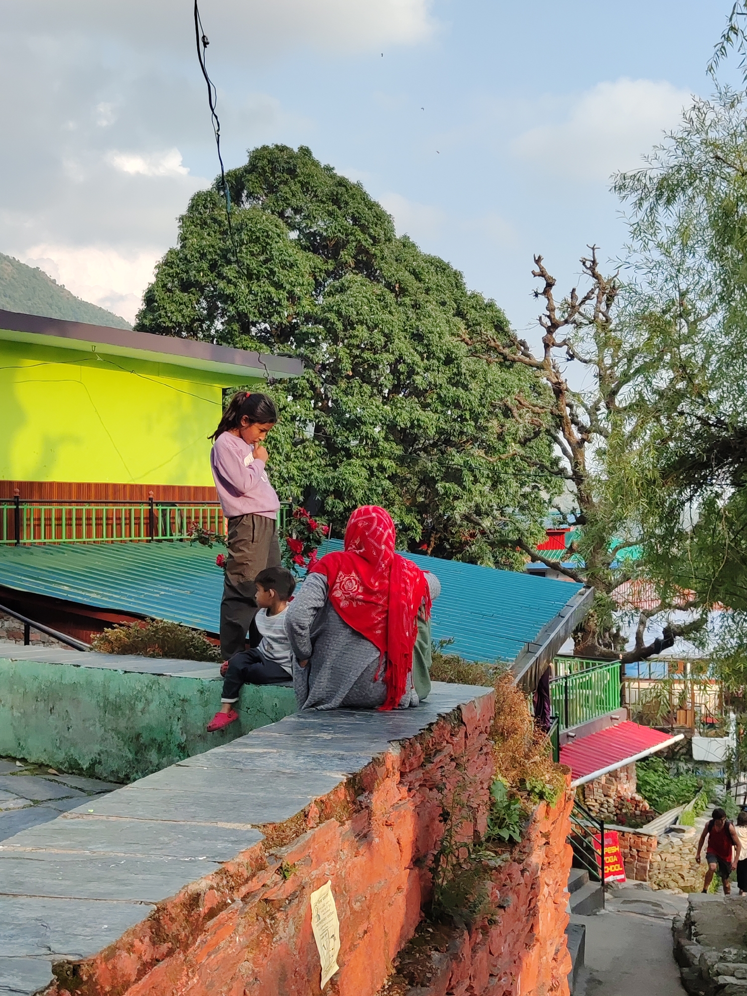 The image depicts an outdoor scene in what appears to be a hilly region, possibly in or near the city of Haini, India, although there is no direct evidence to support this assertion. Three people are sitting or standing on a low stone wall. A young girl in a pink sweater and dark pants is standing at the edge of the wall with her arms crossed, seemingly pensive. Beside her, a young boy is seated. An adult, possibly a woman, is seated next to the boy, wearing a grey outfit and a red headscarf. The wall is composed of stone and brick, with a mixture of green plants growing on the surface. In the background, there are buildings with roofs of green, blue and red and exteriors of bright lime green color with green railings. Behind the buildings, a large, full tree dominates the landscape. To the right, there are more trees and foliage. The sky is partly cloudy. In the lower right of the image, a path leads downhill with two people walking. One is wearing a red tank top, and the sign behind the path reads "Fresh Yoga School". The overall setting suggests a residential area with a mix of natural and built environments.