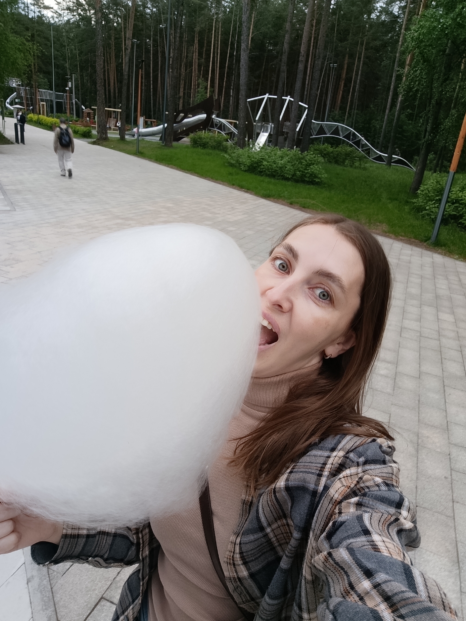 A woman is holding a large white cotton candy in front of her face, with her mouth open as if to take a bite. She has brown hair, fair skin, and is wearing a beige turtleneck sweater and a plaid flannel shirt. She is standing on a gray-paved walkway, with trees and playground equipment visible in the background. The photograph appears to be taken in the city of Perm, Russia, as the background matches common sights in the city. Two other people are walking further down the walkway.