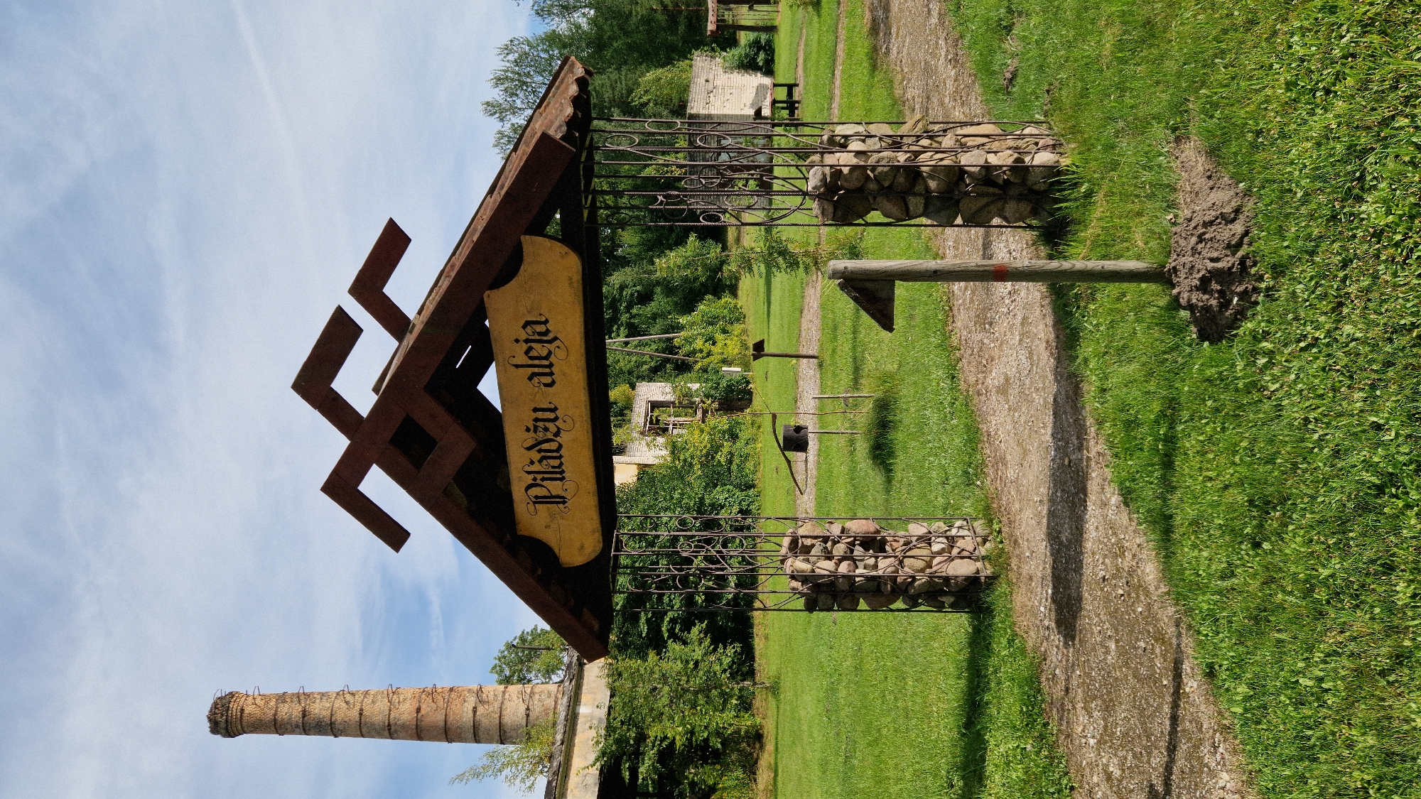 In Kazdanga, Latvia, an entrance archway stands on a gravel path in a grassy field. The archway has a wooden roof with a decorative metal frame. A yellow wooden sign with the inscription "Pilādu aleja" hangs from the arch. The supports of the archway are constructed from metal cages filled with stones. To the left of the archway, a tall brick chimney rises against a partly cloudy sky. Behind the archway, several dilapidated brick buildings are visible, partially obscured by dense green foliage. The ground is covered in a mix of green grass and a gravel path. Several small, wooden posts are scattered throughout the area.