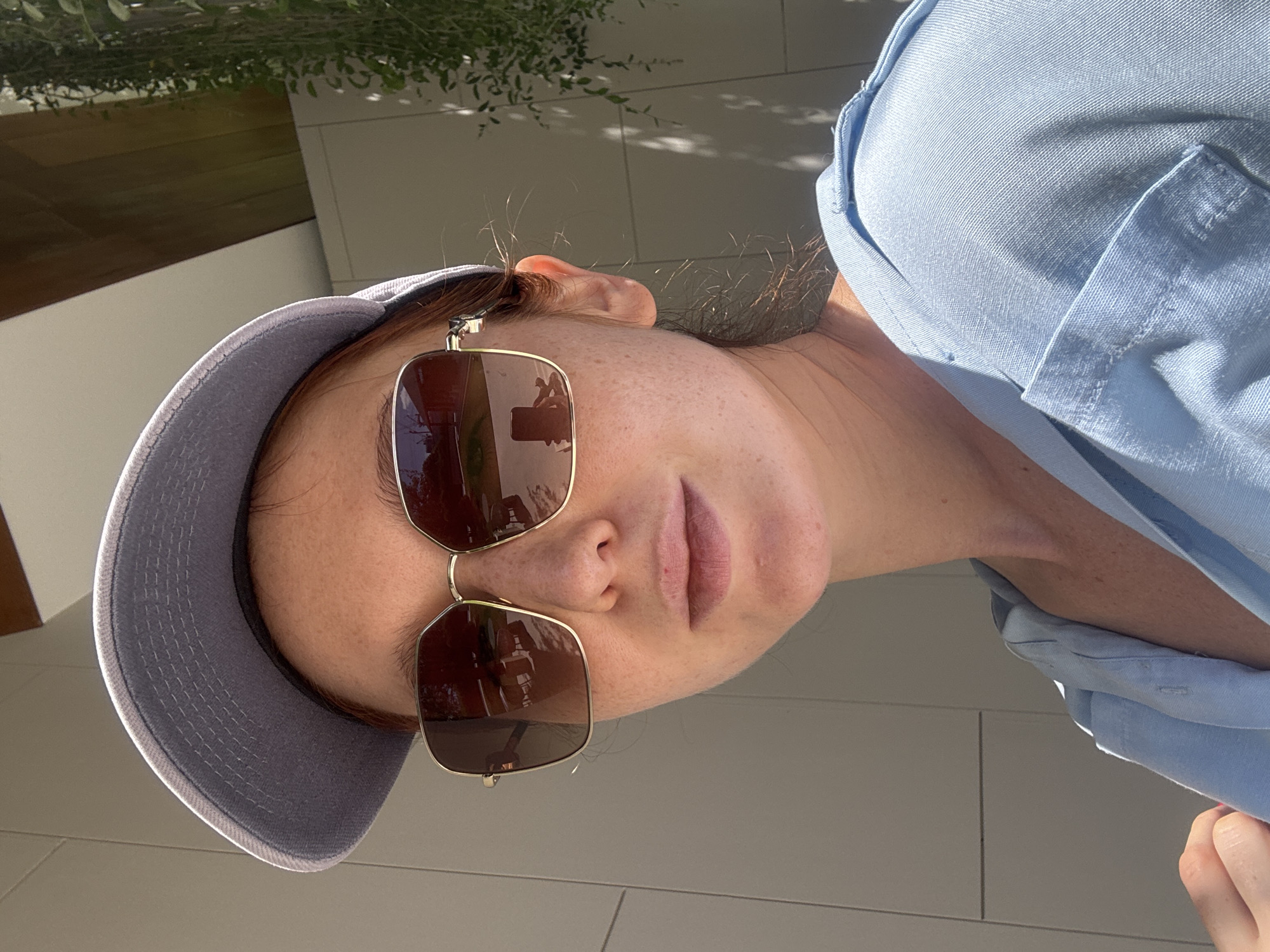 A woman wearing a blue baseball cap and brown-tinted sunglasses is seen from a close-up, slightly angled perspective. She is wearing a light blue collared shirt. The background includes light-colored surfaces that appear to be part of a building or architectural structure, with some glimpses of greenery. Given the prompt's inclusion of Choeng Thale, Thailand, it's reasonable to infer the photo was taken in this location, although there are no specific landmarks to confirm it from the image alone.