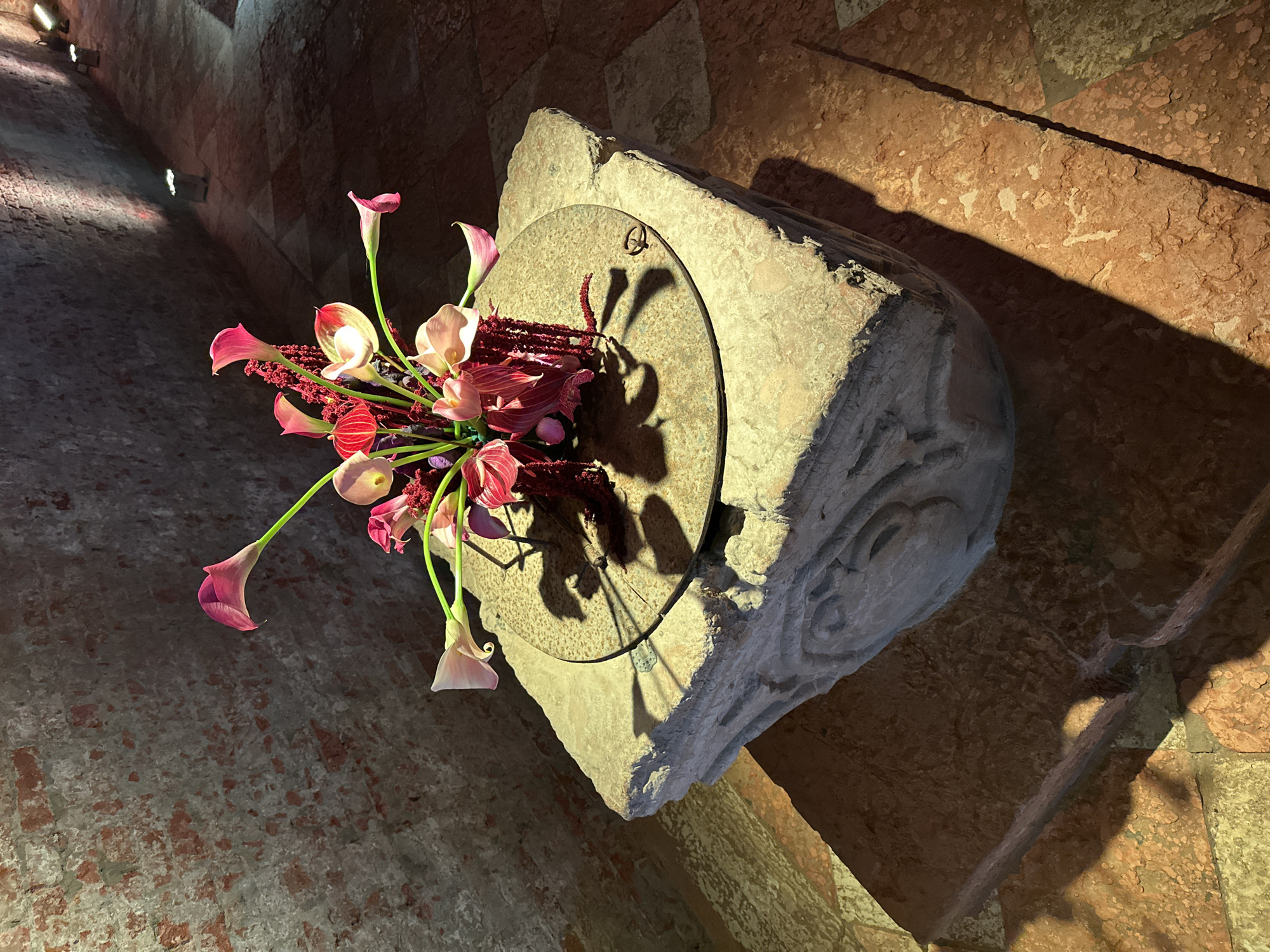 The image captures an artistic arrangement in what appears to be an interior space of a building in Venice, Italy. The primary subject is a stone structure, possibly a fragment of an architectural element, adorned with a floral arrangement featuring pink and white calla lilies and other red and dark-colored flowers. The stone has a circular inset on its surface, which casts a shadow resembling the flowers' form. The walls in the background show Venetian brickwork with visible wear and age. Light sources are positioned overhead, creating highlights and shadows that accentuate the textures of the stone and the walls.