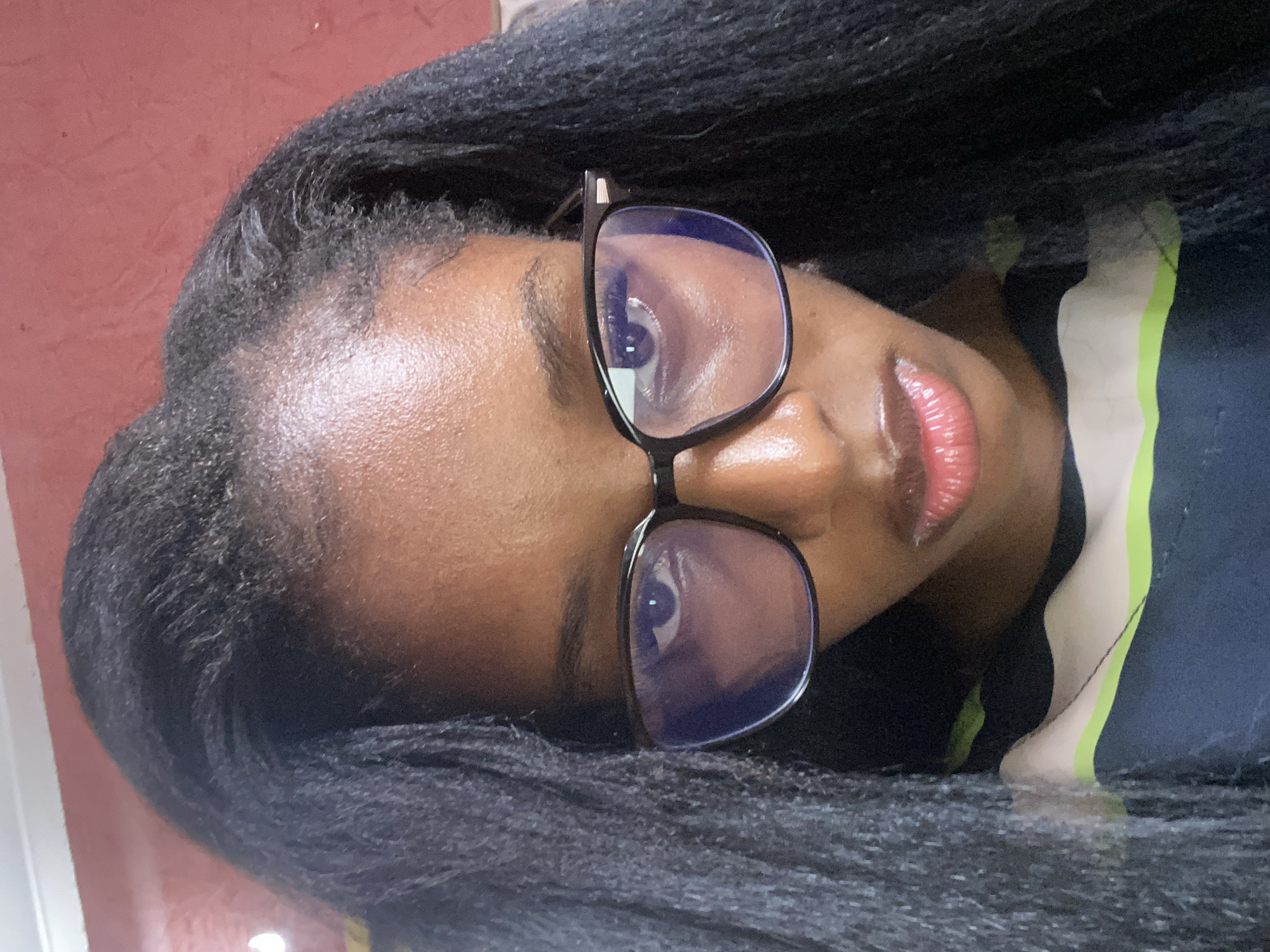 A close-up, front-facing, over-the-shoulder shot features a young Black woman wearing black-rimmed glasses and a multi-colored striped shirt. The woman has dark skin and her dark, braided hair is draped over her shoulders and frames her face. The background is a plain, reddish-brown wall with a white shelf or trim visible at the top. The lighting suggests an indoor setting, possibly a home. The image appears to have been taken in Jos, Nigeria.
