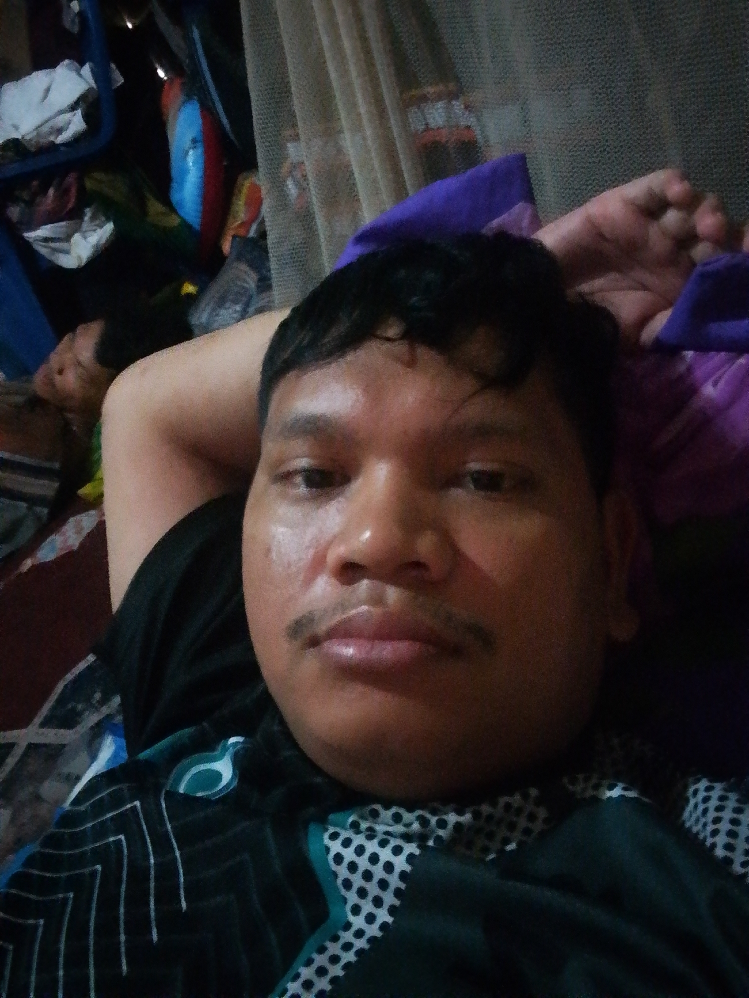 A selfie is taken by a person in a reclined position, likely on a bed. The person is facing the camera with their head resting on their left arm, which is bent and positioned behind their head. The person has dark hair, a mustache, and appears to be an adult male with brown skin. They are wearing a dark t-shirt with a geometric pattern in white and teal. In the background, to the left, another person, possibly a child, is visible lying down and seemingly asleep, with their head partially obscured. The environment appears to be indoors, with a cluttered background that includes what looks like laundry, bags, and possibly some brightly colored inflatable objects. A sheer curtain is visible on the right side of the image. The lighting suggests it might be evening or a dimly lit room. The specific location within Labuhan Bilik, Indonesia, cannot be determined from the visual information alone.