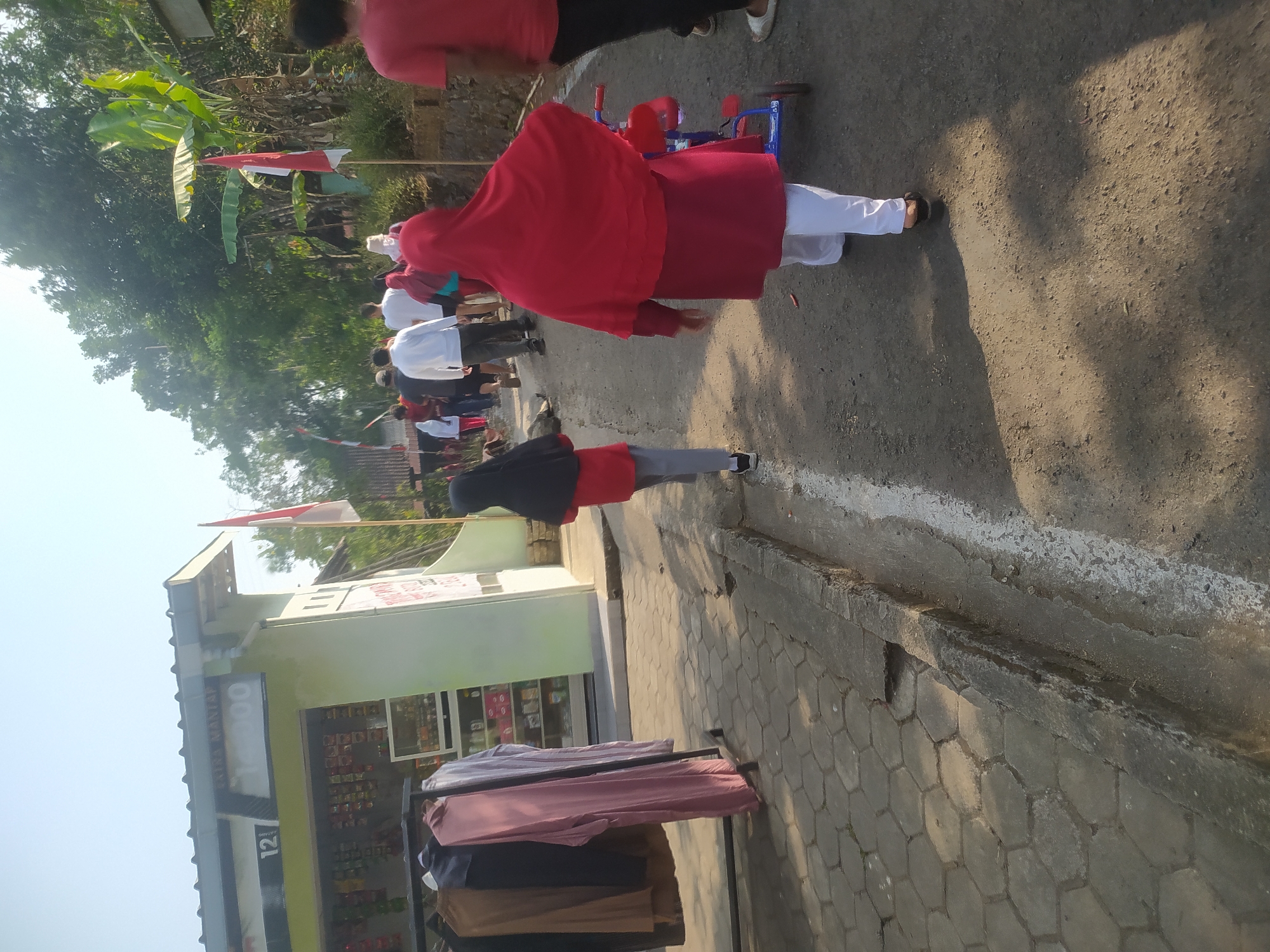 The image depicts a street scene in Lopait, Indonesia, on a bright, sunny day. A group of people, including adults and children, are walking along a paved road lined with hexagonal stones. Many are wearing red clothing, and some are wearing hijabs. In the background, a small green building with a white roof appears to be a shop, with merchandise visible through its windows. A rack of clothing is displayed outside the shop. Indonesian flags are visible, suggesting a national event or celebration. The road has a white line painted on its edge, and shadows from trees and structures are cast across the scene. A small red and blue child's tricycle is being pushed by one of the people. The overall atmosphere seems to be casual and communal, possibly during a morning walk or a local gathering.