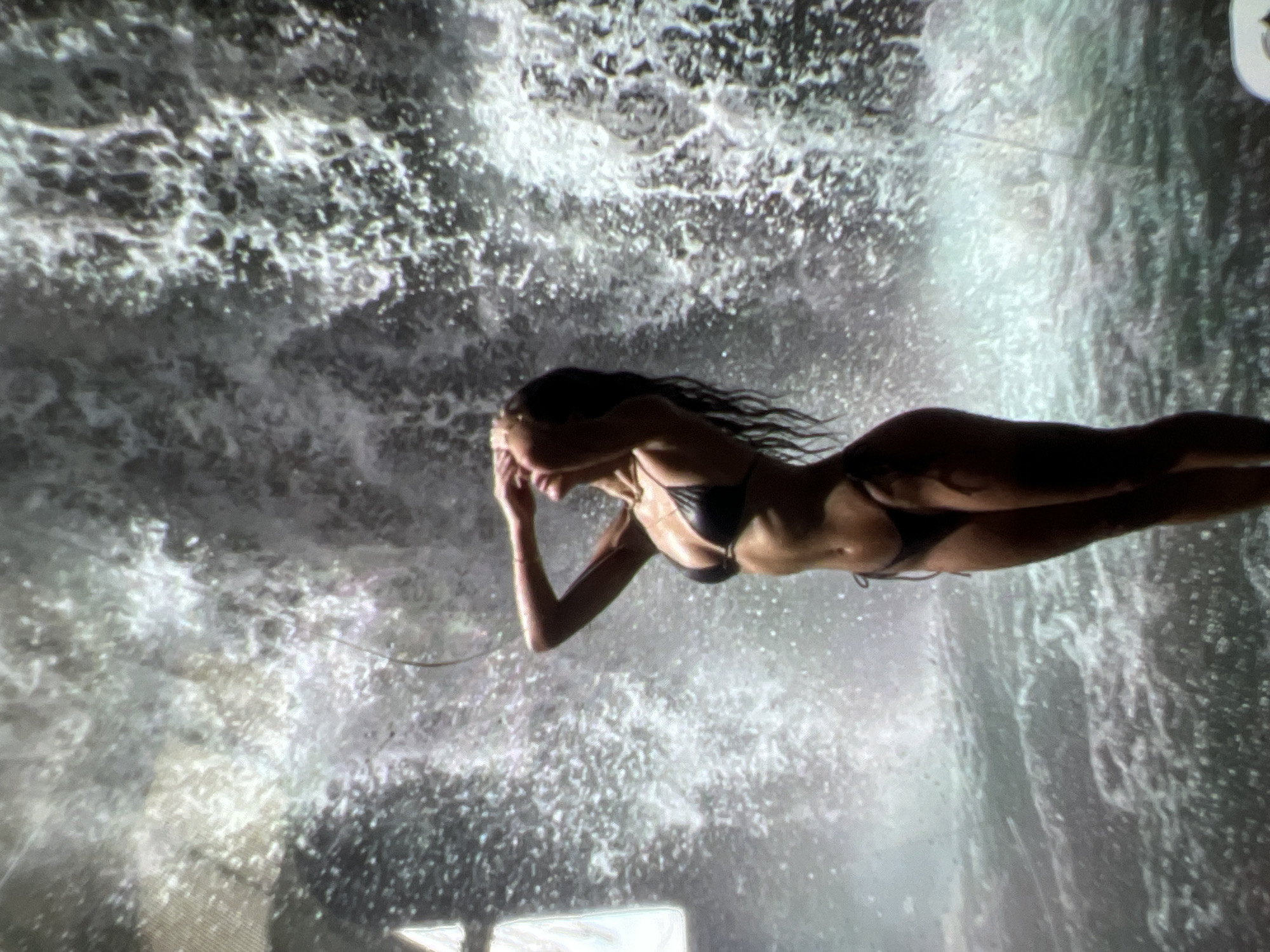 The image shows a woman in a black bikini standing in front of a waterfall. She has her arms raised and her hands are covering her face. The waterfall is a large curtain of water, and the water is splashing around her. The woman's body is toned and slender, and she has long, dark hair that is wet and wavy. The scene is dimly lit, and the water appears to be illuminated by artificial light. The location in the city of Kara-Dıykan, Kyrgyzstan is not explicitly depicted in the image, as the background is a projected image of a waterfall.