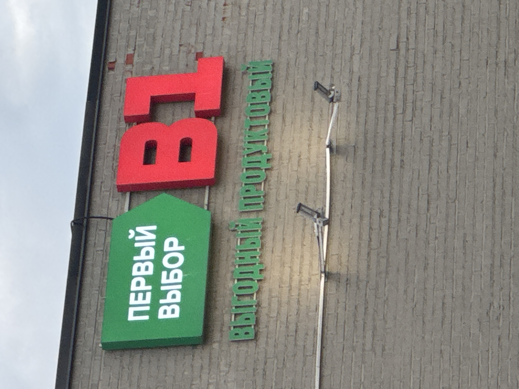 A sign for a B1 grocery store is mounted on a brick wall. The sign is green and displays the words "ПЕРВЫЙ ВЫБОР" in white. To the right of this sign are large red letters that spell out "B1". Below the main sign, smaller green letters on the brick wall spell out "ВЫГОДНЫЙ ПРОДУКТОВЫЙ". There is a security camera mounted on the wall below the signs. The sky is overcast. The store is located in Dedovsk, Russia.