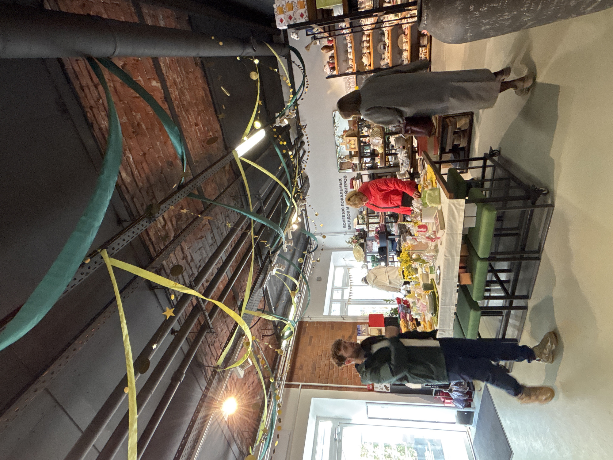 An interior view depicts a brightly lit concept store or market within a renovated industrial-style building, characteristic of creative clusters found in cities like Saint Petersburg, Russia. The ceiling is exposed, featuring black-painted beams, pipes, and original red brickwork, adorned with festive yellow and teal fabric ribbons, gold paper circles, and star cutouts. White walls flank the exposed brick, with a sign on the back wall in Russian reading "БОЛЕЕ 300 ЛОКАЛЬНЫХ БРЕНДОВ И ДИЗАЙНЕРОВ" (MORE THAN 300 LOCAL BRANDS AND DESIGNERS).
Three individuals are visible. In the left foreground, a man with light brown, curly hair and a beard, wearing a dark green jacket, white top, dark trousers, and light brown sneakers, walks towards the right with his left foot in motion. To his right, a long rectangular display table draped with a white tablecloth, holding various items including a vase of yellow flowers, green placemats, and white dishes, is surrounded by green-upholstered stools. Behind this table, a woman with blonde hair, dressed in a bright red long-sleeved top, stands facing and interacting with items on the table. In the right midground, a second woman, seen from behind, walks towards the right, wearing a long grey coat and light-colored chunky sneakers, carrying a dark red shoulder bag. To the far right, a large, dark, textured ceramic pot or urn stands partially visible. Various shelves and display units filled with ceramics, packaged goods, and other merchandise are arranged in the background. Natural light enters through large white-framed doors and windows on the left.