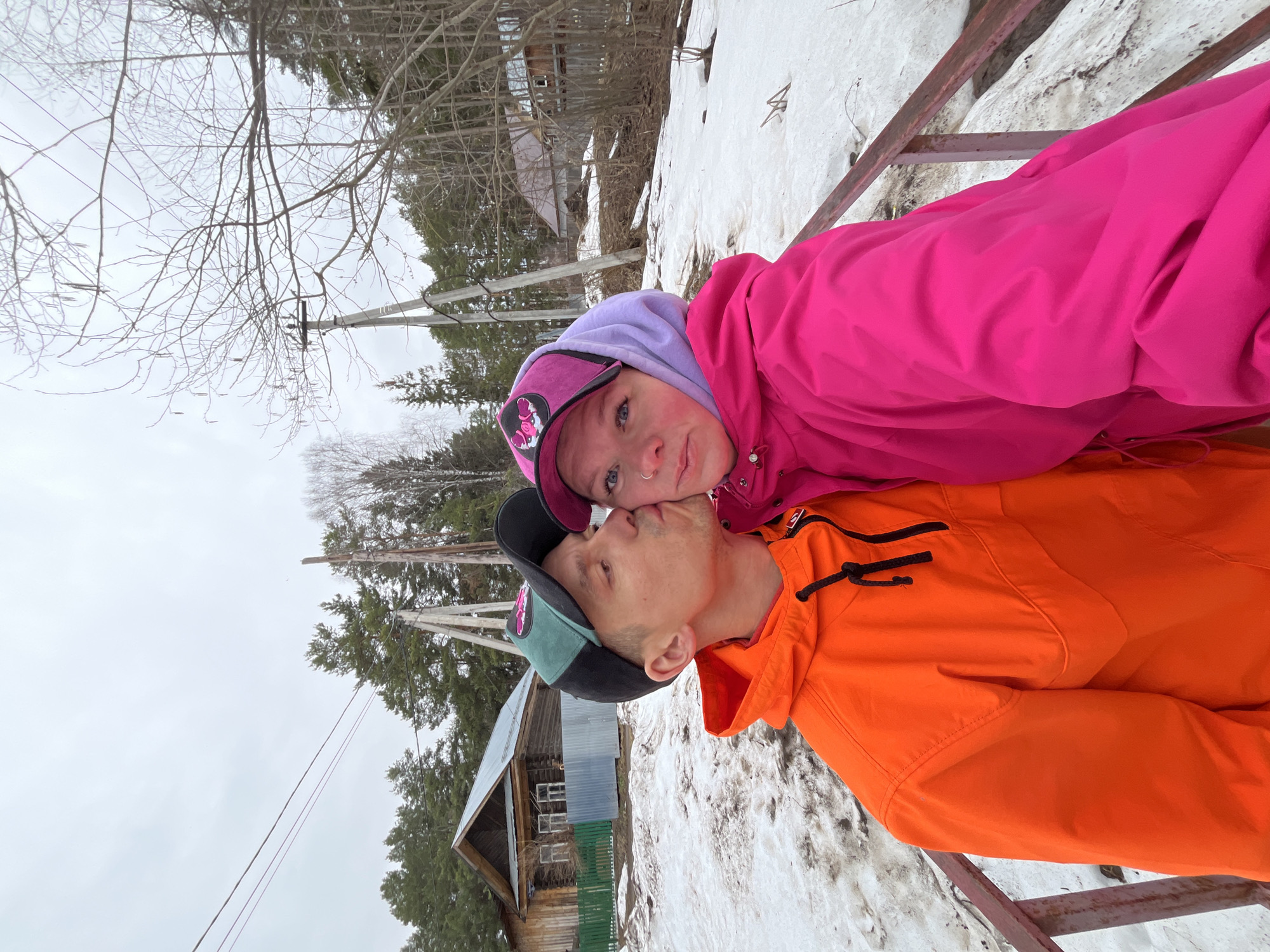 A couple is standing on a metal structure covered in snow, possibly a staircase or a bridge, with a partially obscured wooden building and a snowy field in the background. The woman, wearing a pink jacket and a purple hoodie with a cap, is looking at the camera. The man, wearing an orange jacket and a cap, is turned slightly to the side and appears to be about to kiss or is kissing the woman's cheek. The setting appears to be in a snowy, wooded area on an overcast day. Utility poles and bare trees are visible in the background. The location may be in a rural area in the city of Chaikovsky, Russia, as indicated.