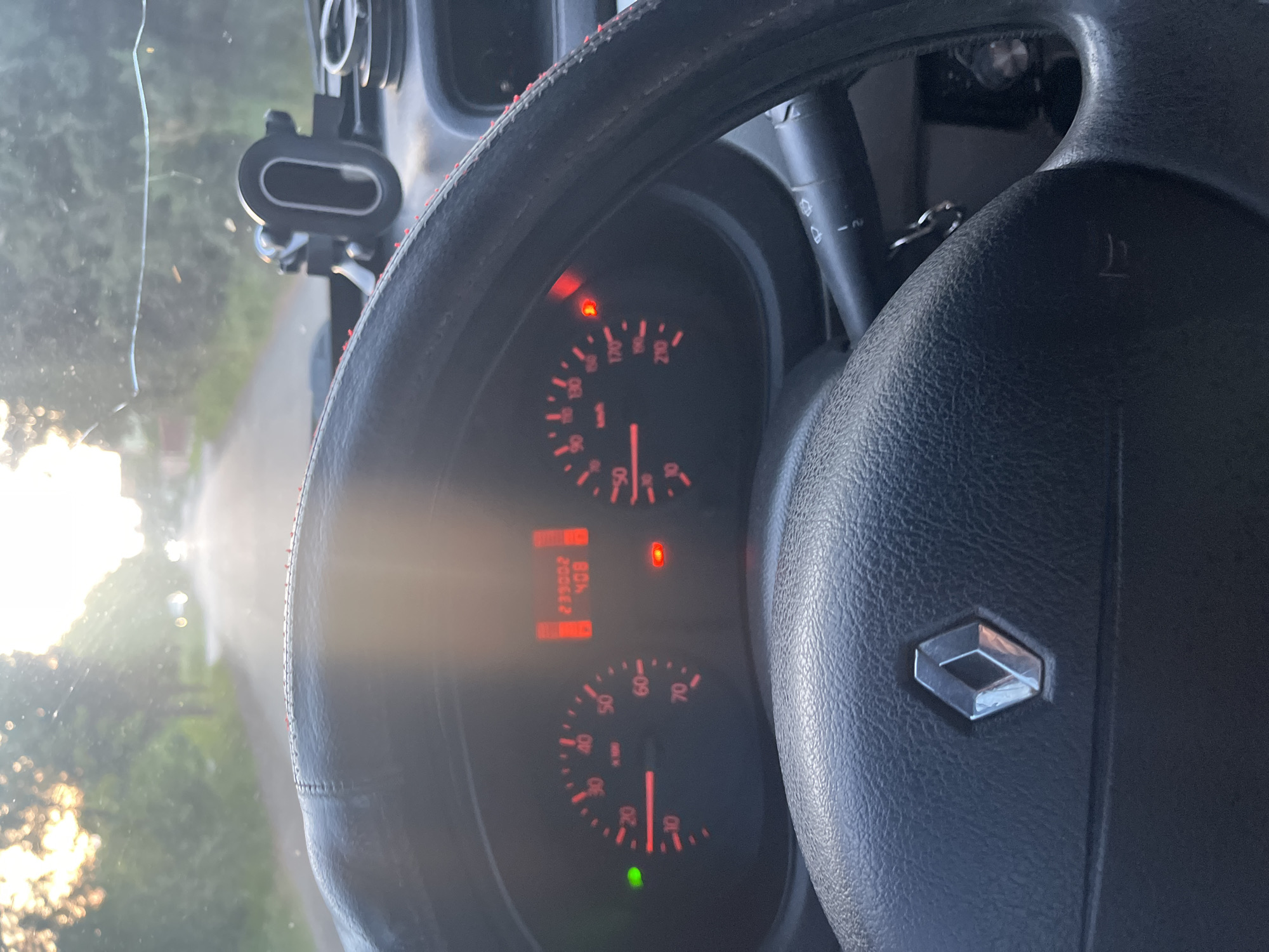 This image captures the interior of a car from the driver's perspective, looking out through the windshield. The scene is heavily influenced by bright, low sunlight streaming in from the left, creating lens flare and a washed-out effect on the left side of the frame.

The main subjects are the car's steering wheel and dashboard. The black steering wheel, partially covered with a red-stitched rim, dominates the right and upper portions of the image. A metallic silver Renault logo is centered on the steering wheel hub. To the left of the steering wheel, a control stalk with symbols for wipers and their speed settings (0, 1, 2) is visible.

The dashboard instruments are illuminated, displaying a speedometer showing 0 km/h and a tachometer showing 0 RPM, indicating the car is stationary with the engine off or idling. A digital display shows an odometer reading of "235552 KM" and the text "40B". Several warning lights are active: a red light resembling an oil lamp or engine fault indicator is illuminated, along with another red warning light above the tachometer. A green light is also visible at the bottom of the speedometer, likely for parking or low beam lights.

Through the windshield, a paved road is visible, flanked by green trees and foliage. A thin scratch or streak is present on the windshield on the upper left. A black phone holder or mount is attached to the upper right of the windshield. A key chain with keys hangs from the ignition, partially visible between the steering wheel and the dashboard.

The low angle of the sun suggests either early morning or late afternoon/evening. The weather is clear and sunny. The location appears to be a rural or suburban road in Storozhnytsia, Ukraine. No people are visible.