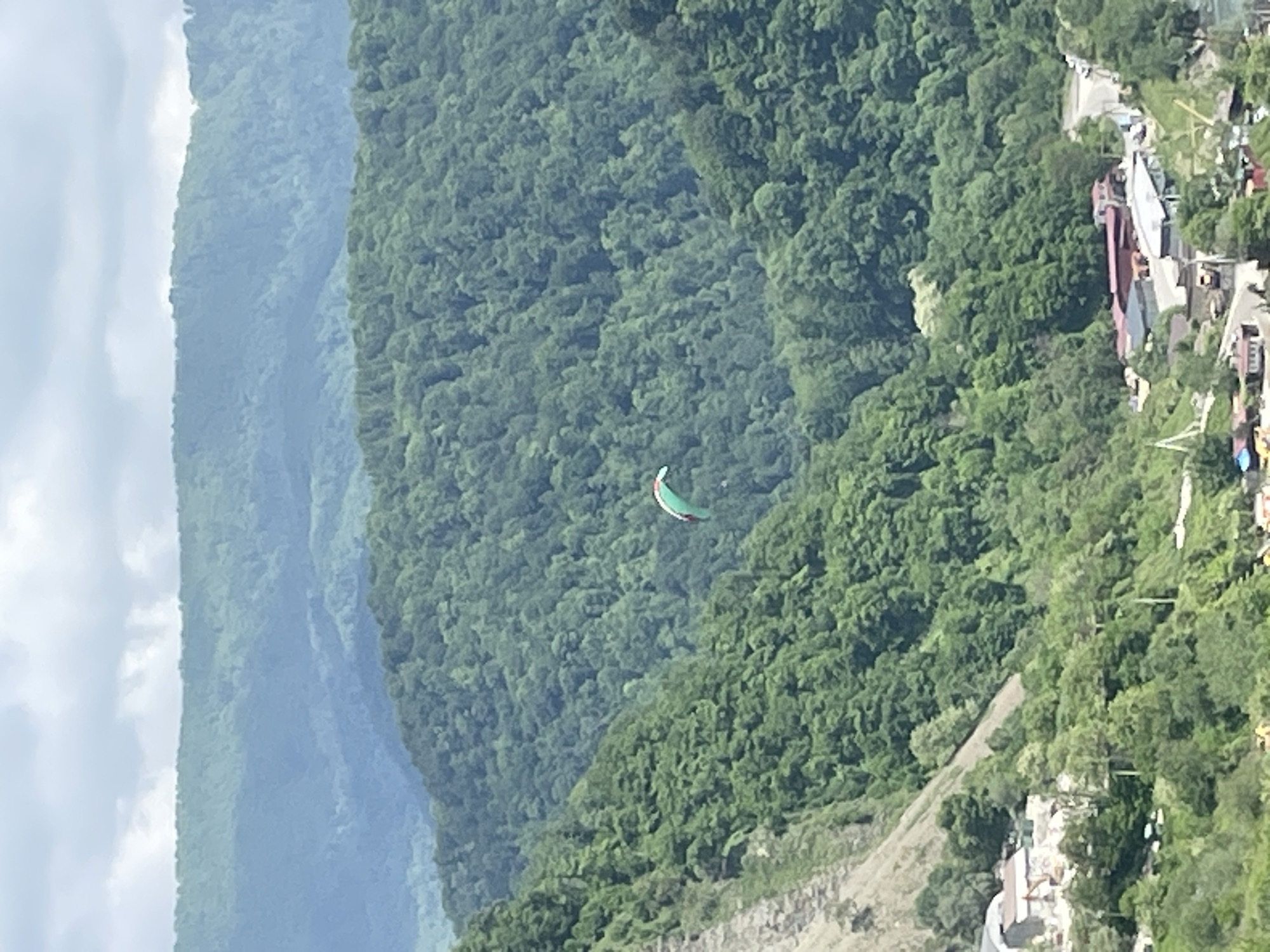 The image shows a paraglider in flight over a densely forested area. The paraglider is positioned in the middle of the frame, with lush green trees filling the majority of the background. In the distance, there are mountainous terrains covered with trees and cloud cover. On the right side of the frame, a small settlement with buildings and a road can be observed. The overall environment suggests a natural, mountainous landscape with a recreational activity taking place. Based on the prompt, the location is near or within the city of Baranovka, Russia.