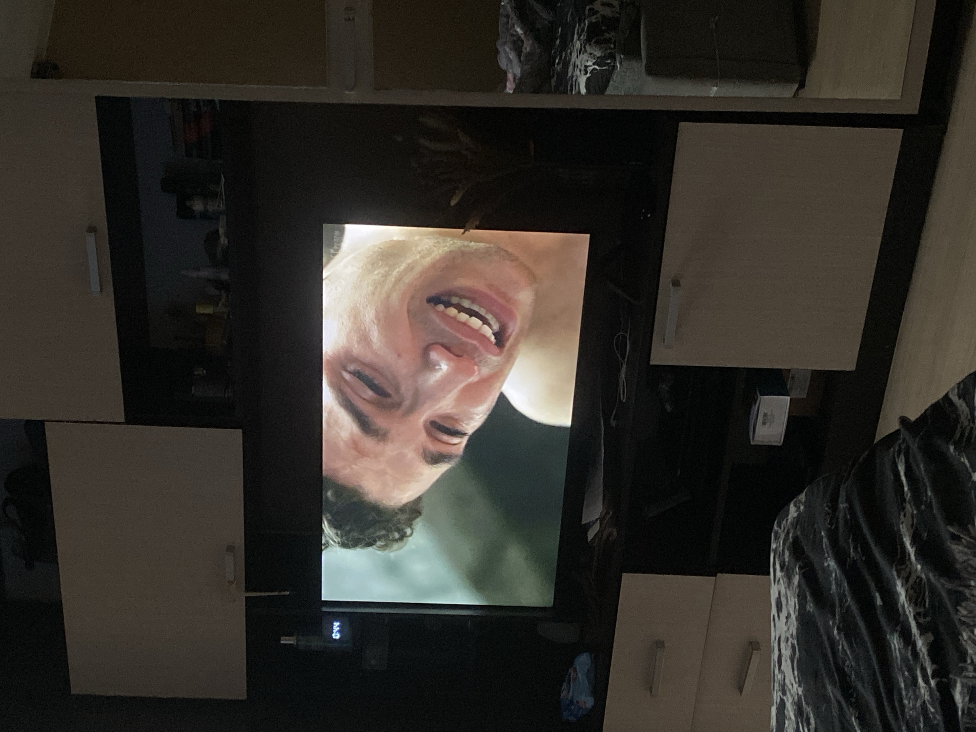 A television screen displays a close-up of a man's face, twisted in an expression of distress or pain. The TV is positioned against a background of kitchen cabinets with simple, light-colored doors. Other items are visible on the shelves above the cabinets. To the right, a patterned piece of clothing or material is partially visible. The location is an interior space, likely a home. There is no information about the location in the city of Yekaterinburg, Russia.