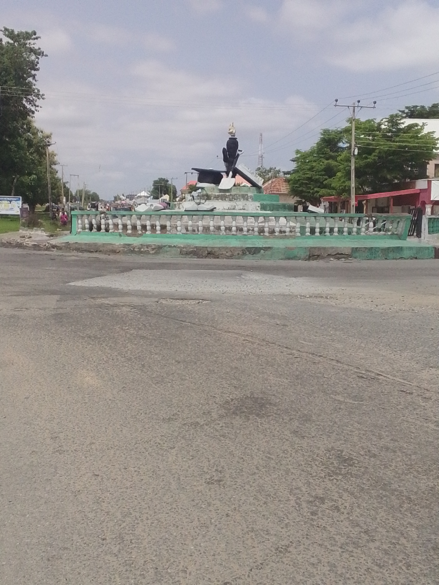 The image shows a monument in Jalingo, Nigeria. The monument is a sculpture of a person in black clothing, holding a shovel, with a golden crown on its head. It is situated on a raised platform with a green base and a white decorative railing. The monument is surrounded by a circular pavement. In the background, there are trees, buildings with red roofs, and utility poles. The sky is cloudy. A road is visible in the foreground, with some cracks and potholes. A few people are walking near the monument, and a sign board is visible on the left side of the image.