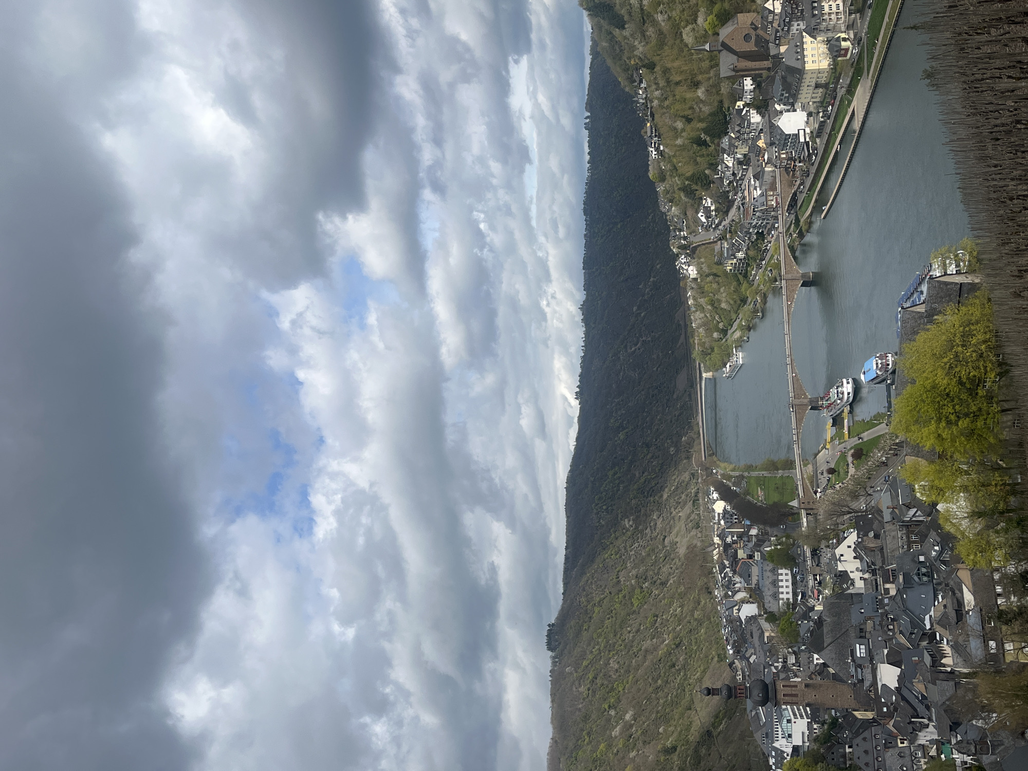 Here's a detailed description of the image:
**Location:** The image shows the city of Cochem, Germany, from an elevated vantage point. The Moselle River flows through the center of the city.
**Environment:** A cloudy sky dominates the left side of the image. The river is flanked by a forested hillside on one side and the city on the other.
**Objects and Subjects:**
* The Moselle River is a central feature.
* A long bridge spans the river.
* The city is comprised of dense clusters of buildings with dark, steeply pitched roofs, including a church.
* Boats are docked along the river's edge.
* Trees and vegetation are interspersed throughout the city and cover the hillside.