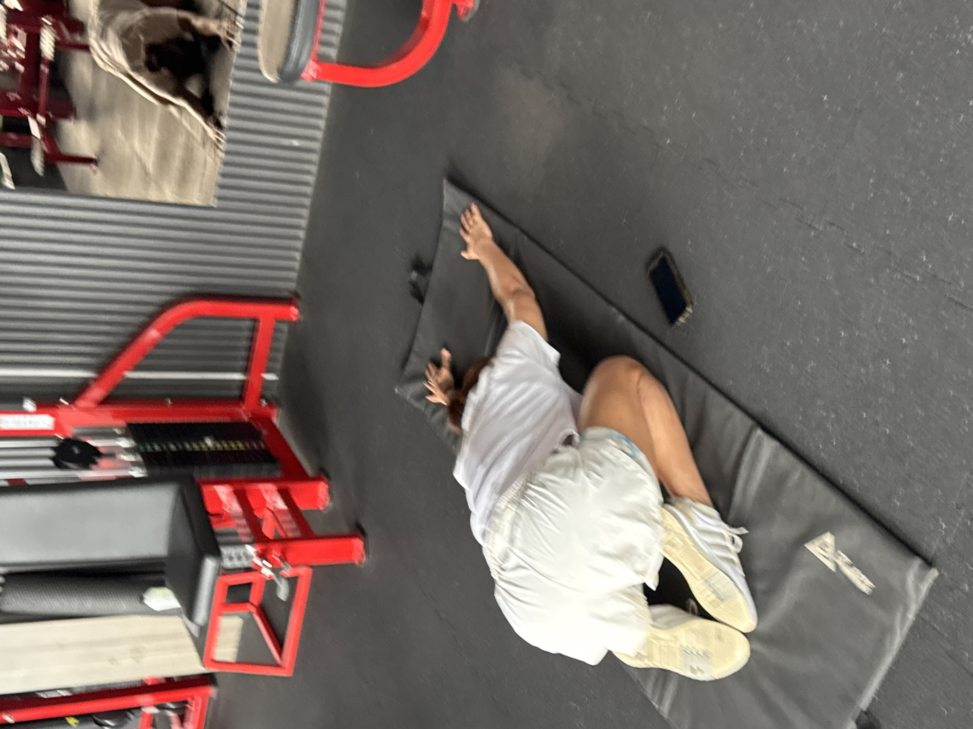 A person is lying face down on a black yoga mat in a gym, performing a stretching exercise. The person is wearing white shorts and a white t-shirt, with white sneakers on their feet. A smartphone is placed on the floor next to the mat. In the background, there are gym equipment with red frames and ribbed metal walls. A reflection in a mirror shows another person or a dog in a similar pose. The gym appears to be located in Kolkata, India.