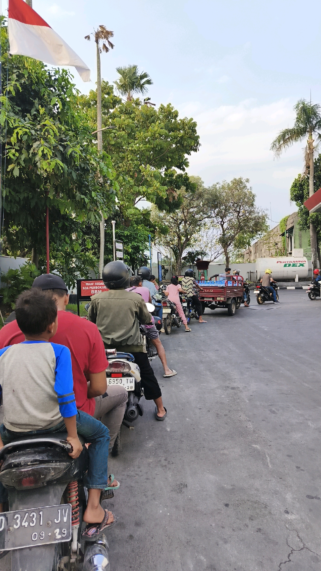 In Karangduren, Indonesia, a line of motorcycles and a small truck are waiting at what appears to be a fuel station or a similar service point. The foreground shows the rear of two motorcycles, one with a young boy in a blue and grey shirt and jeans sitting in front, and another behind him with a person in a red shirt and grey pants. The second motorcycle has a license plate "D 3431 JV 09-29". Behind them, a line of people on motorcycles continues down the asphalt road, which is lined with trees and vegetation on the left. A red and white Indonesian flag is visible flying from a pole on the far left, behind lush green trees. Further down the road, a small red truck is visible, carrying what appears to be bottled water. In the background, a building with a "DEX" logo is present. The sky is overcast.