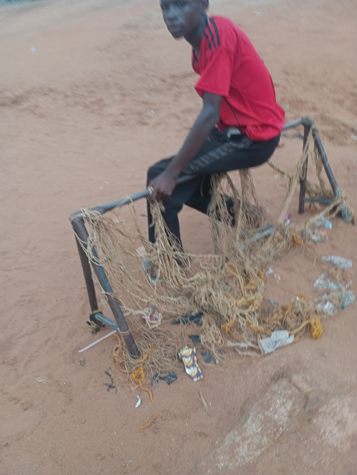 A young person wearing a red shirt with black stripes on the sleeves and black pants is sitting on a makeshift goalpost structure in a sandy area. The goalpost appears to be constructed from metal pipes and is covered with a tangled, worn-out net. Scattered around the base of the goalpost and on the net are various pieces of litter, including plastic bags and bottles. The ground is covered in light brown sand, suggesting an outdoor, possibly rural or informal urban setting. The background is blurred but appears to be a flat, open expanse, consistent with an outdoor location in Onitsha, Nigeria, where such informal sporting setups are common.
